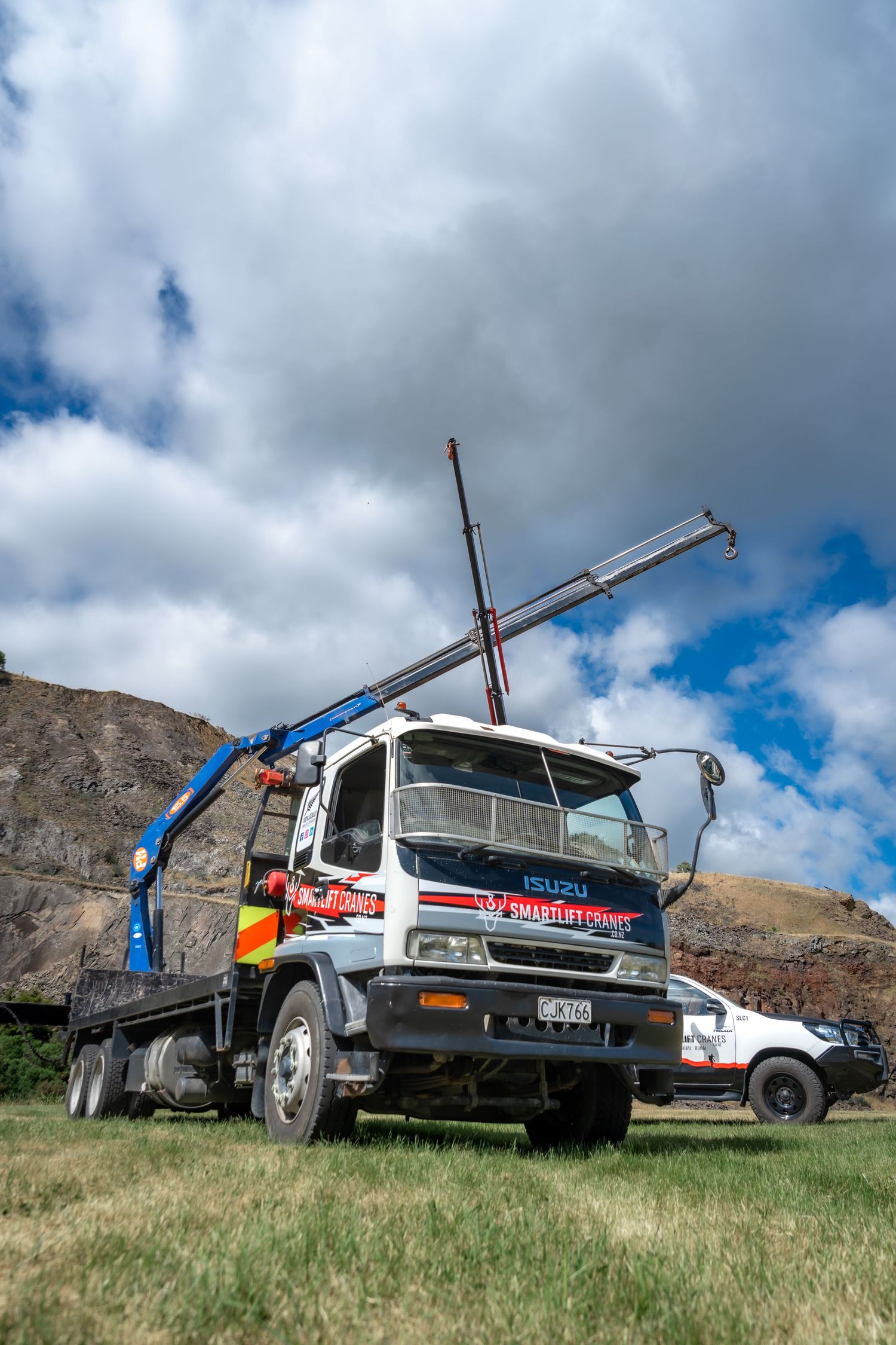 Isuzu crane truck with extended boom arm parked on grass under cloudy sky near rocky hillside.