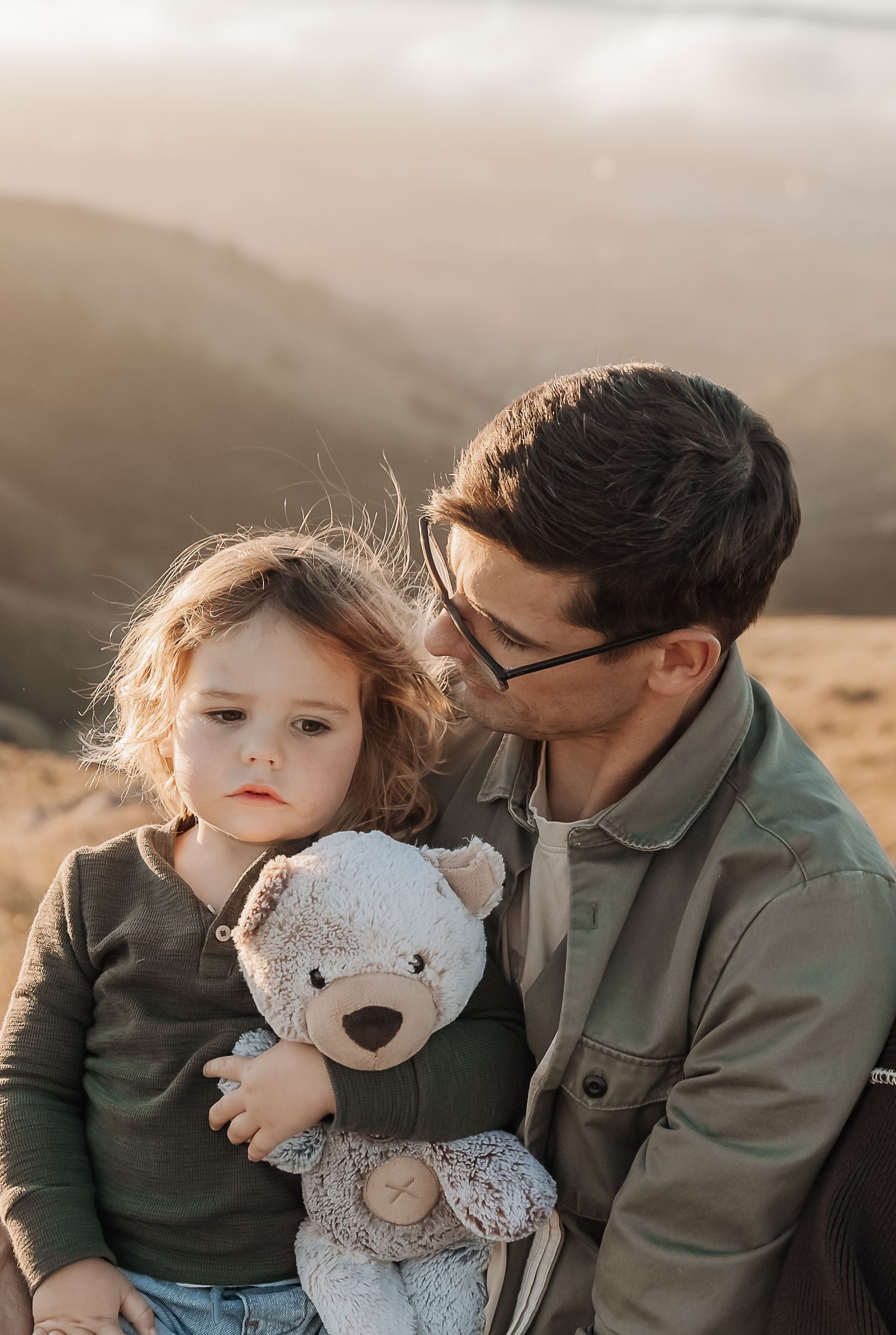 A father tenderly holds his young child clutching a teddy bear outdoors with golden hills in the background.