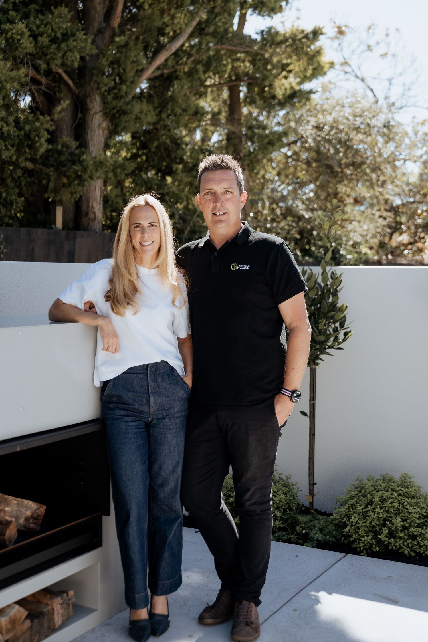 A smiling man and woman pose together outdoors on a modern patio with white walls and lush greenery.