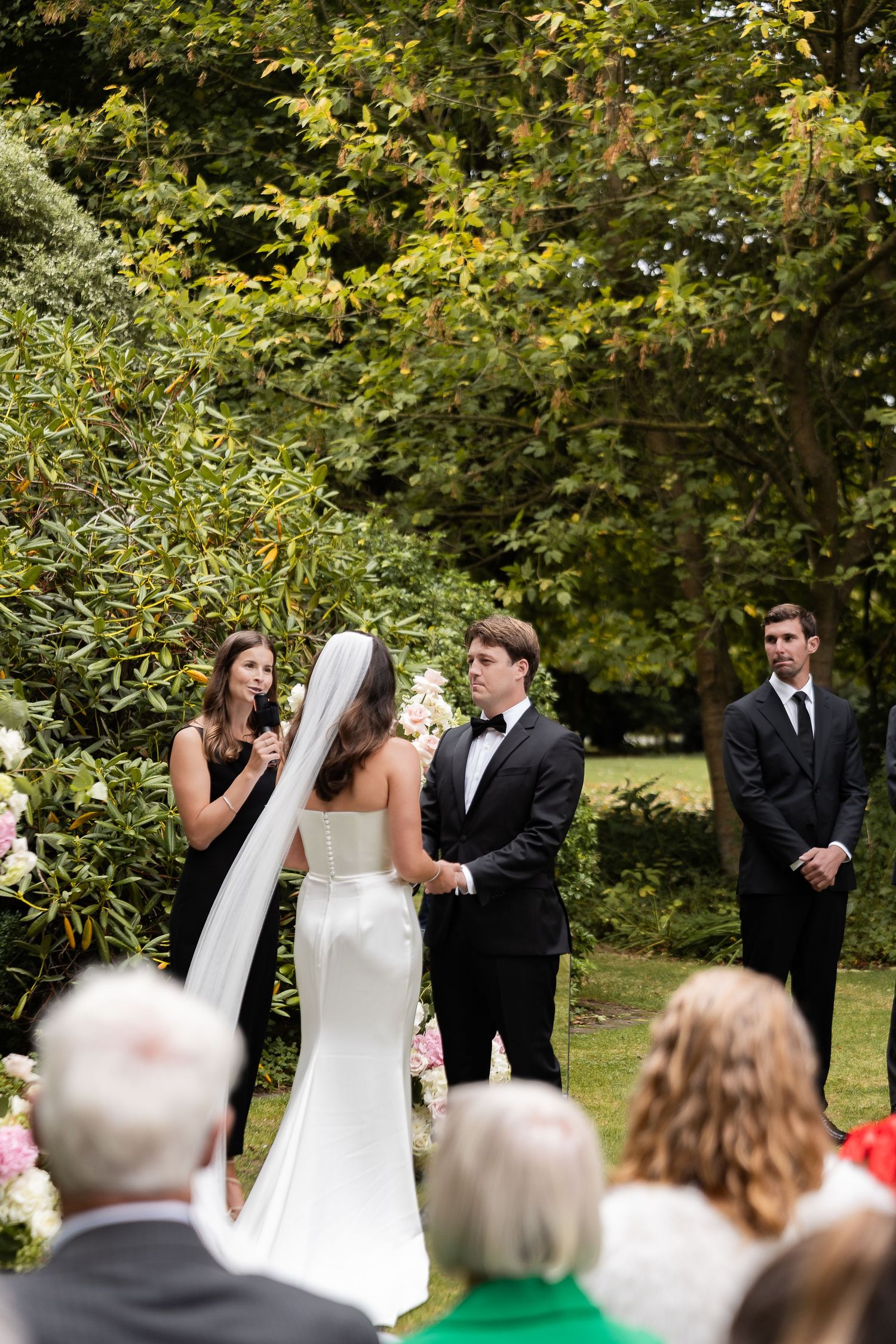 Outdoor wedding ceremony with bride in white gown and veil, groom in tuxedo, officiant, and guests among lush green trees.
