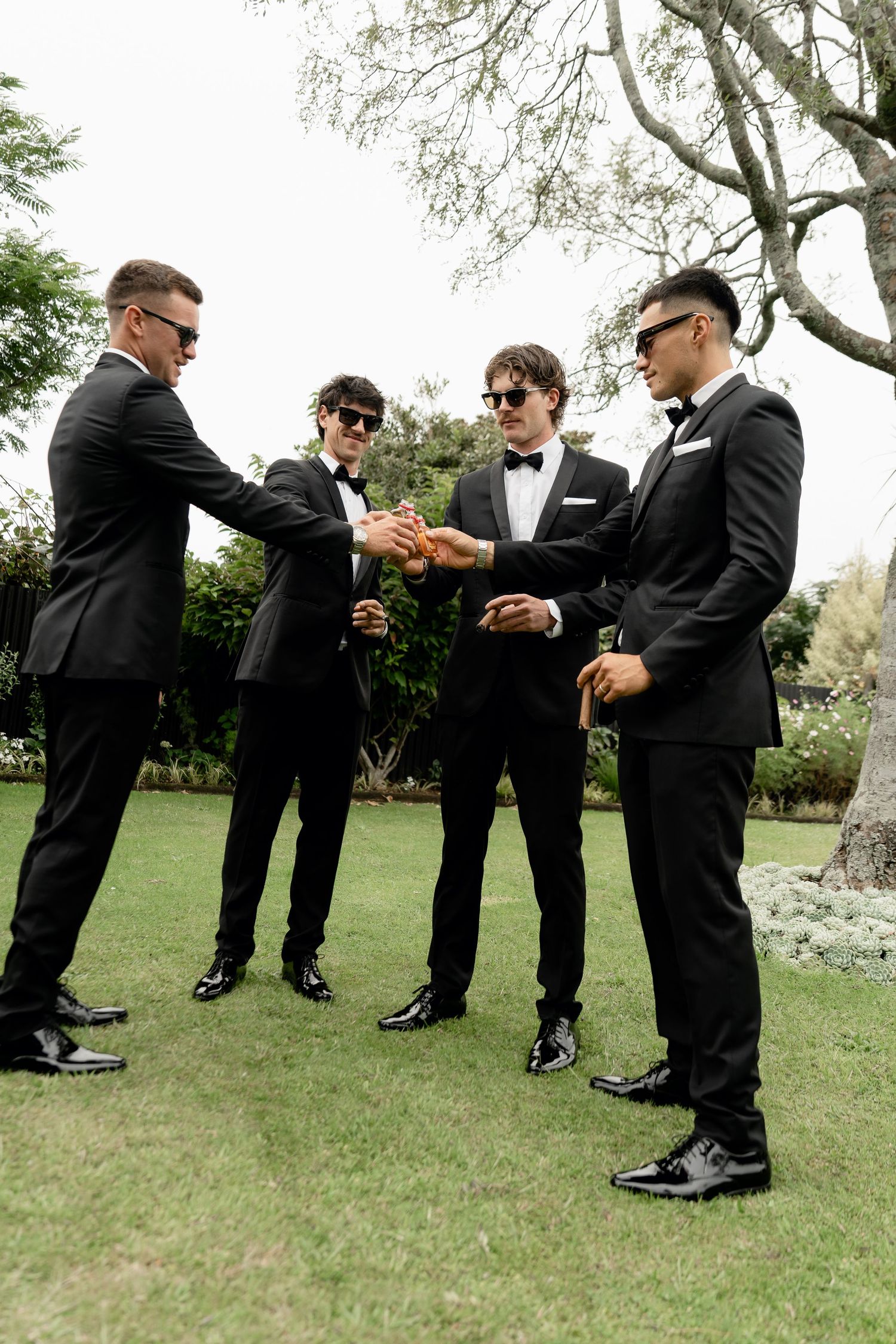 Four groomsmen in black tuxedos and sunglasses gather outdoors, stacking hands together in a celebratory gesture.