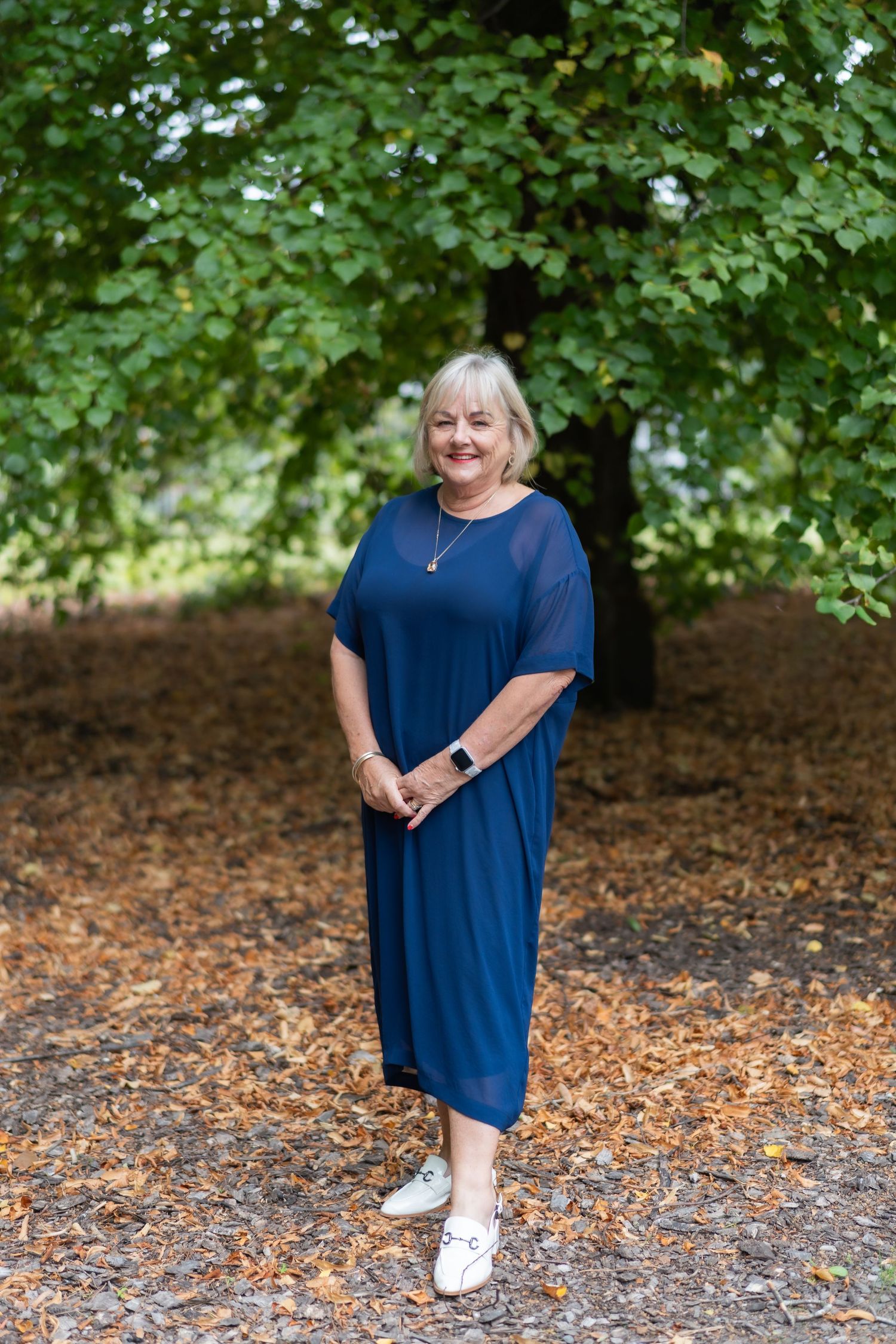 Smiling woman in navy blue midi dress standing among autumn leaves under green trees.
