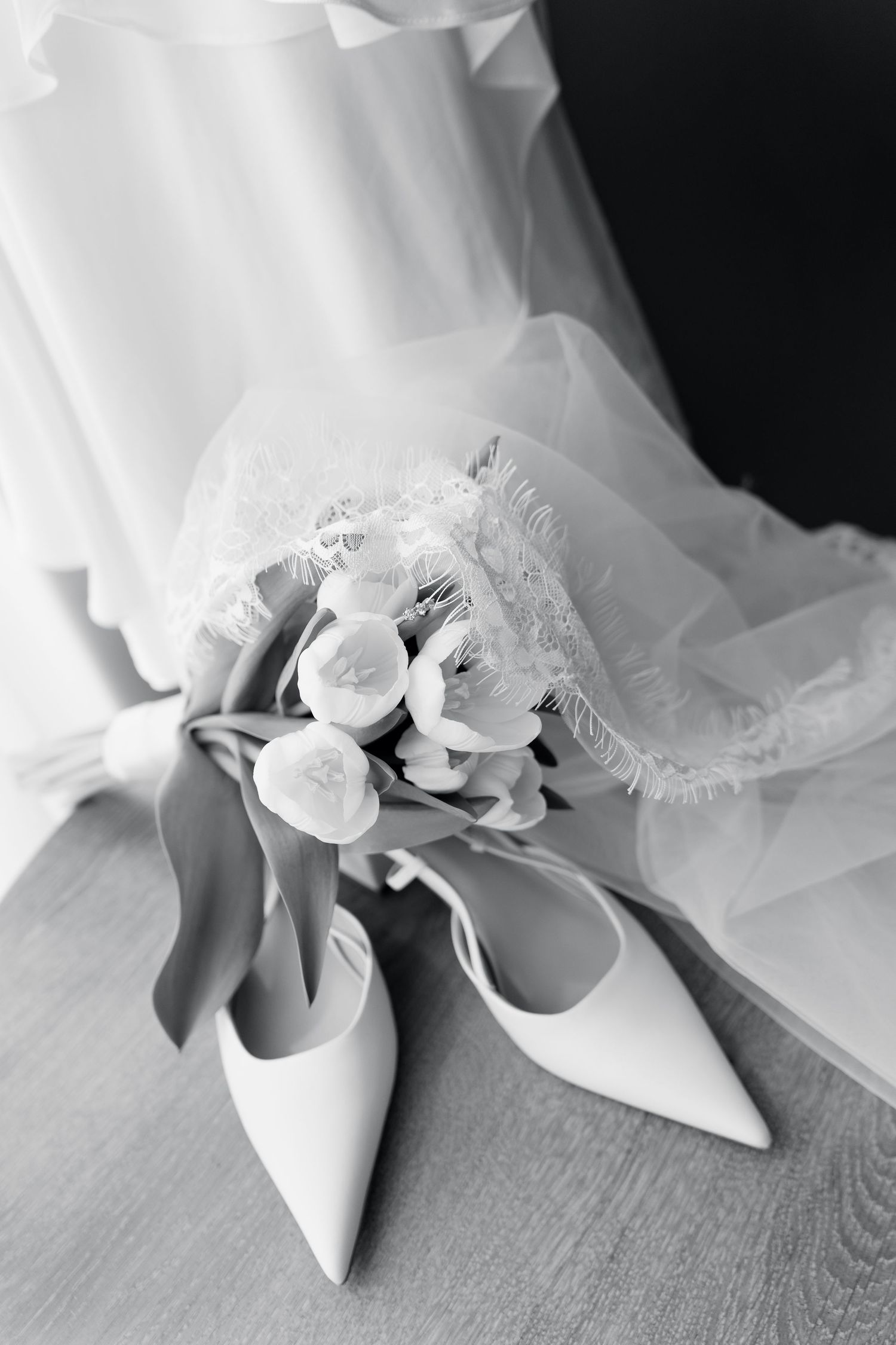 Black and white photo of bridal heels and flower bouquet beside a white wedding dress on a wooden floor.