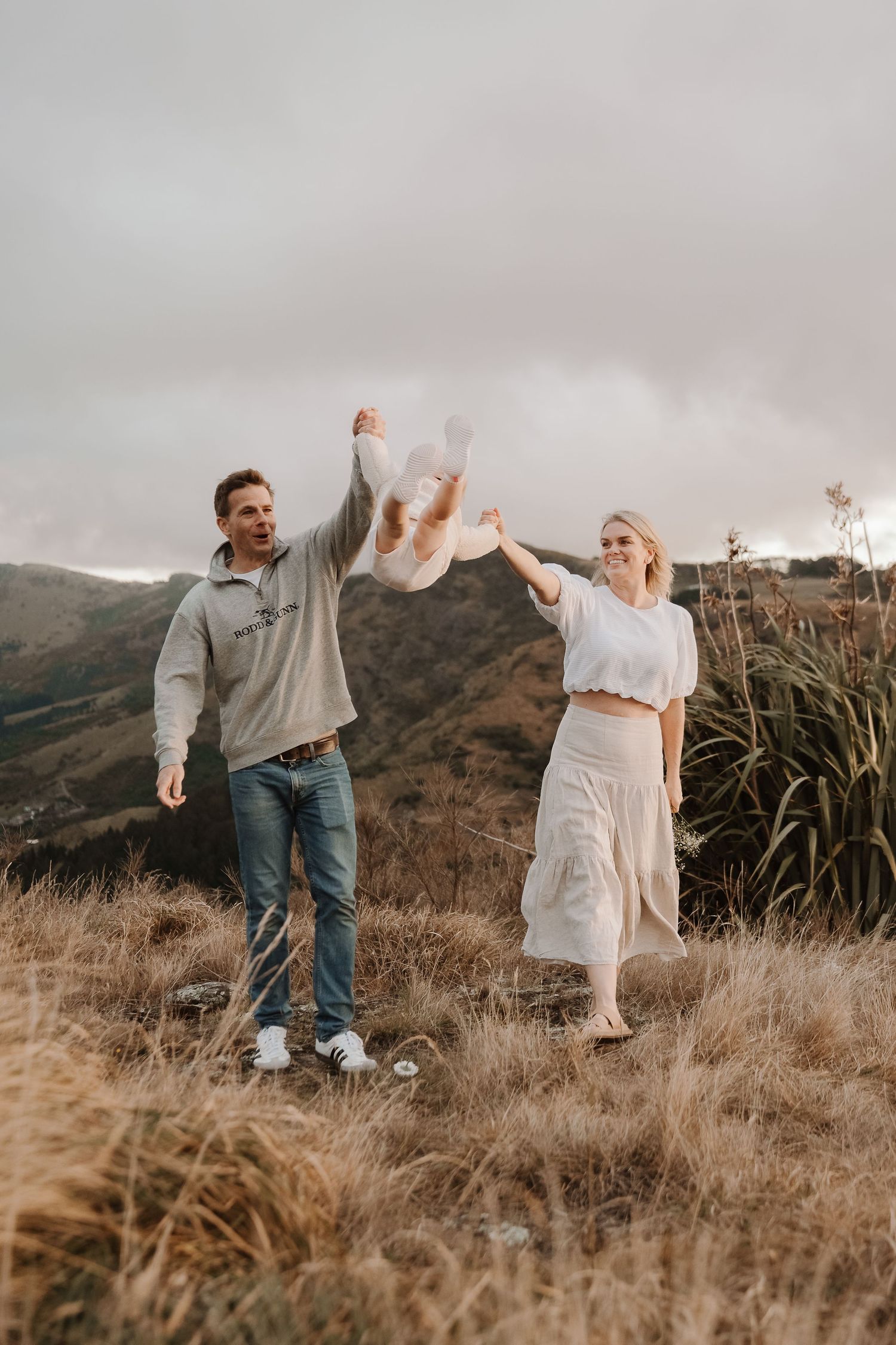 Couple joyfully lifting baby in the air on a scenic hilltop with golden grass and misty mountains.