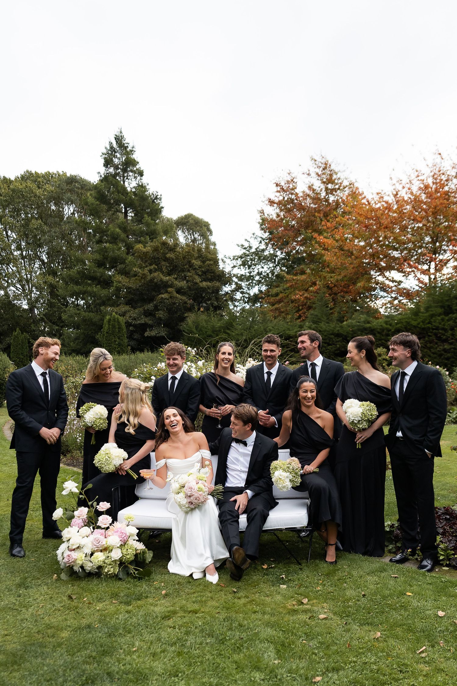 Christchurch wedding party in black attire poses with bride and groom on white bench in lush garden, holding white floral bouquets.
