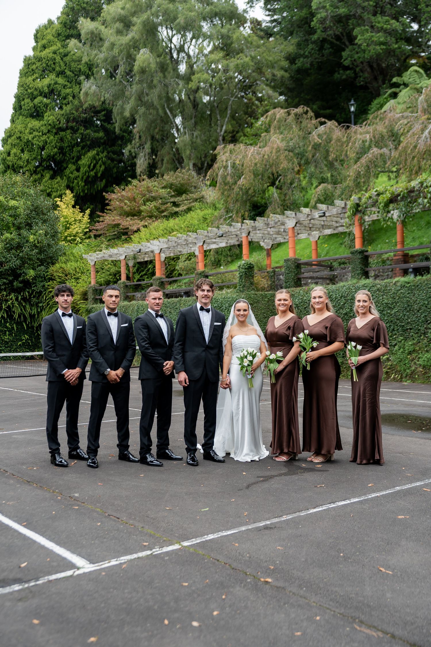 Wedding party poses outdoors, groomsmen in black tuxedos and bridesmaids in brown gowns flank bride and groom.