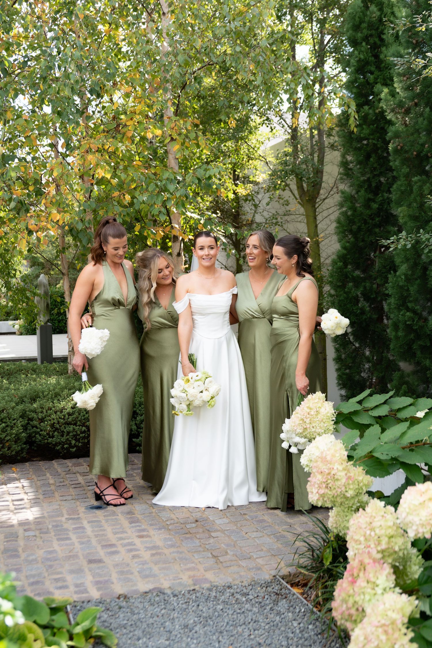 Bride in white gown with four bridesmaids in sage green dresses holding white bouquets in a garden setting.