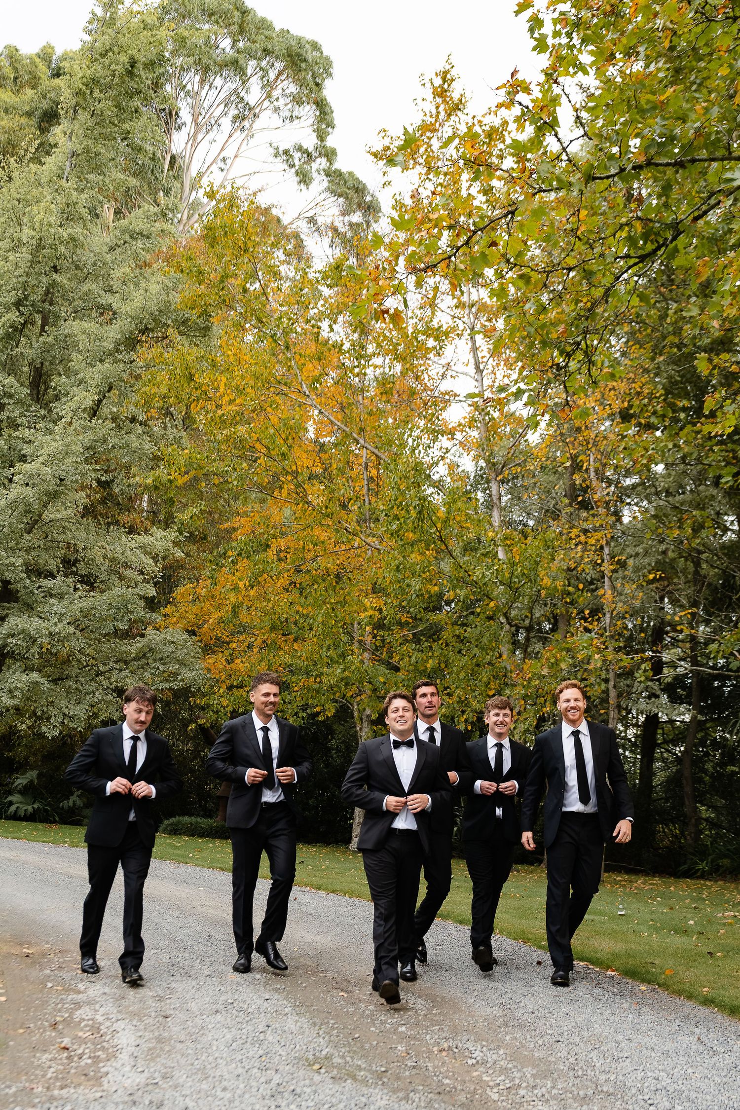 Groom and groomsmen in black suits walking on a gravel path surrounded by autumn trees.