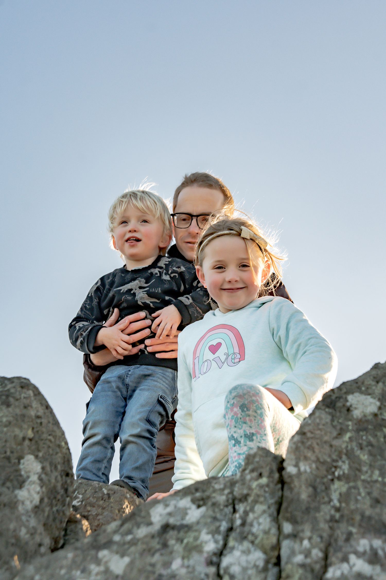 Two young children and their parent sit together on large rocks during golden hour against a bright sky at the Christchurch Port Hills.