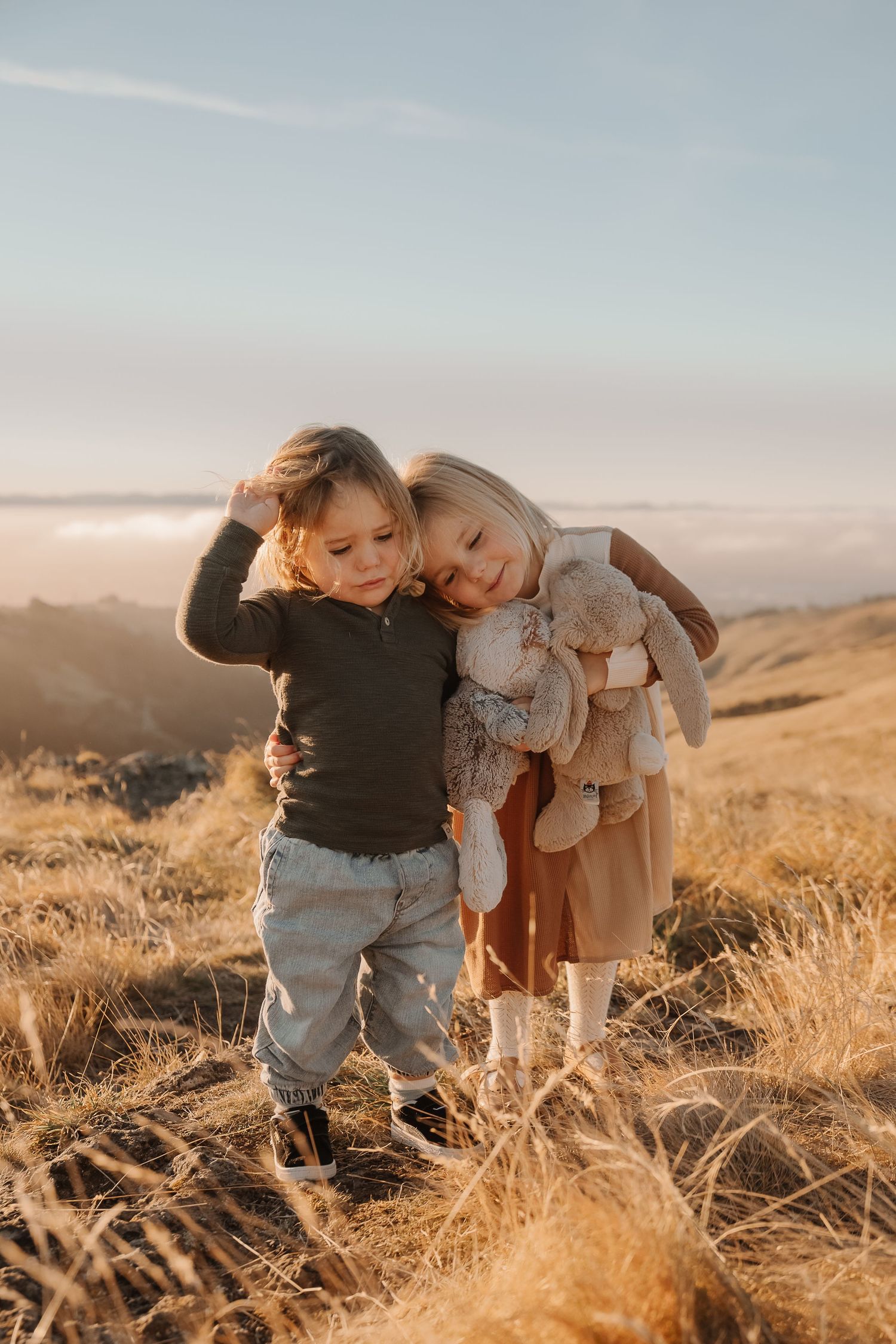 Two young children embrace on a golden hillside at sunset, girl holding stuffed bunny toy.