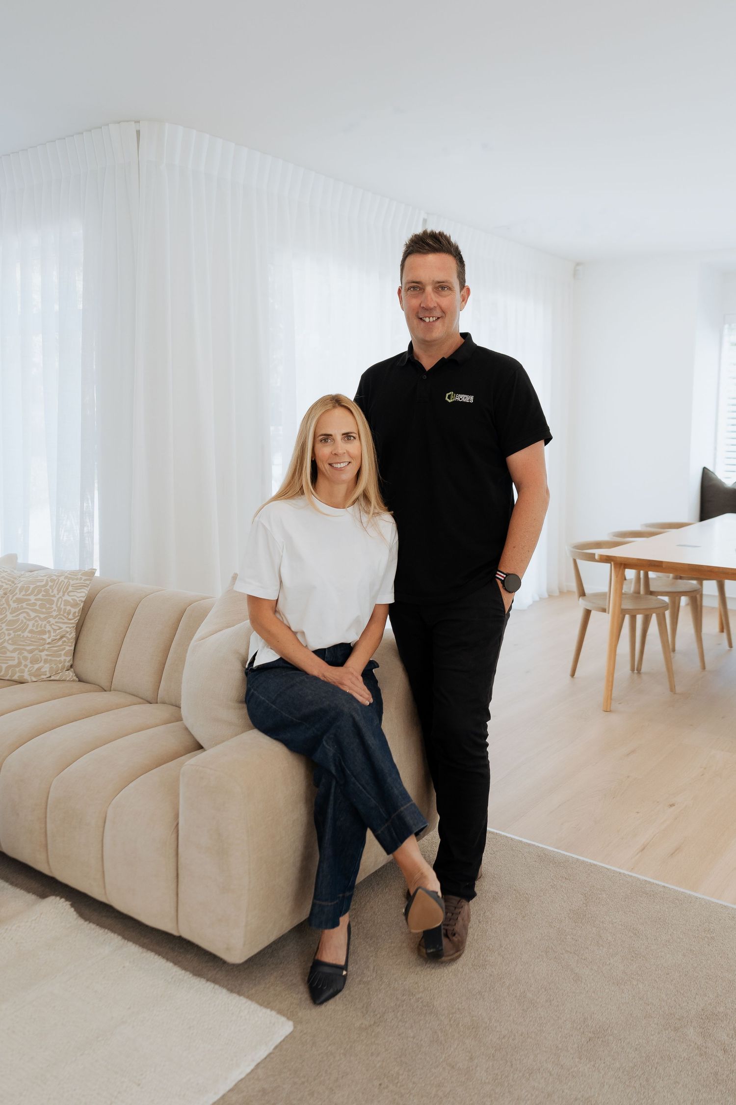 Smiling blonde woman and tall man in black polo posing together in a modern white living room.