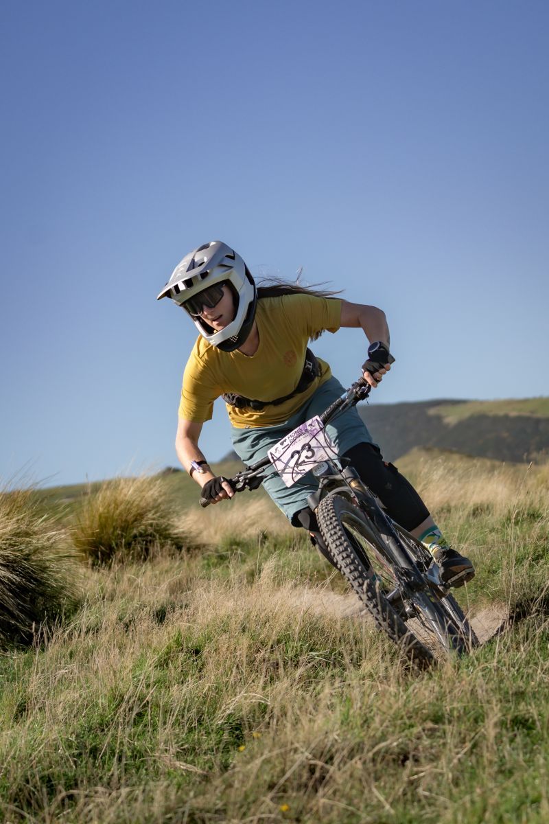 Mountain biker wearing protective gear carves through grassy hillside terrain during downhill ride.
