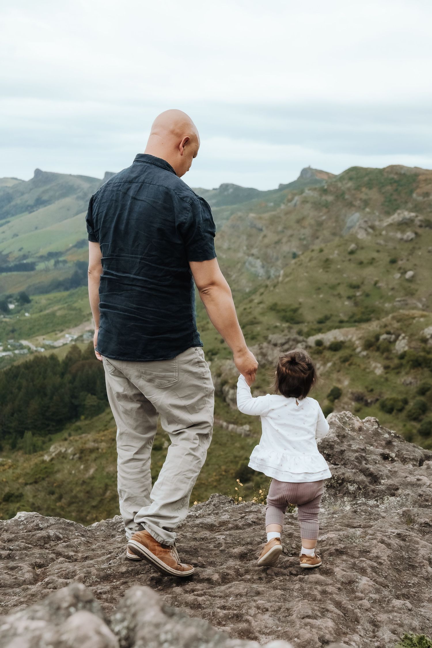 Bald man holding hands with toddler girl on rocky mountain summit, overlooking green valley landscape.