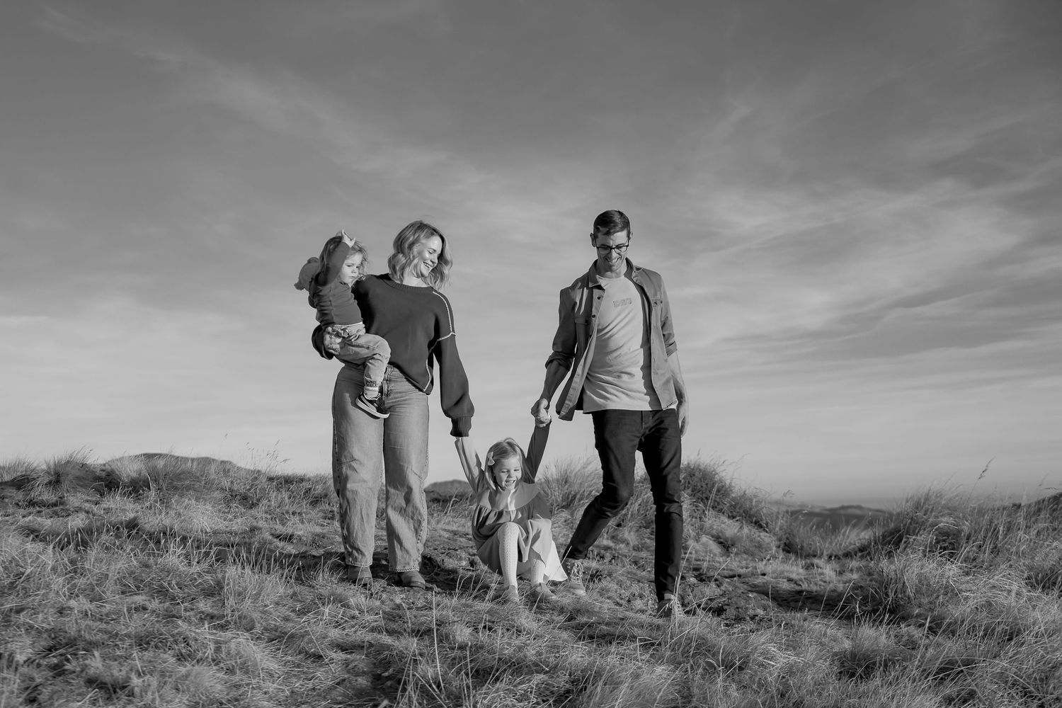 Black and white photo of a happy family walking on a grassy hill under a cloudy sky.