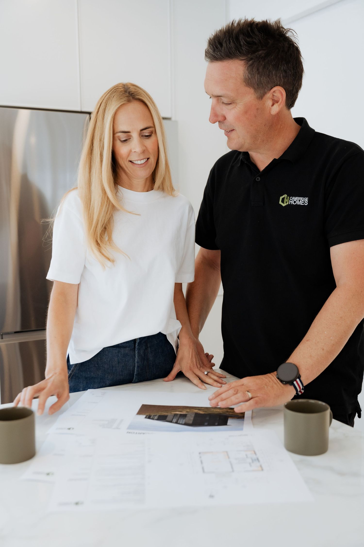 A man and woman review architectural blueprints and material samples together at a white kitchen counter.