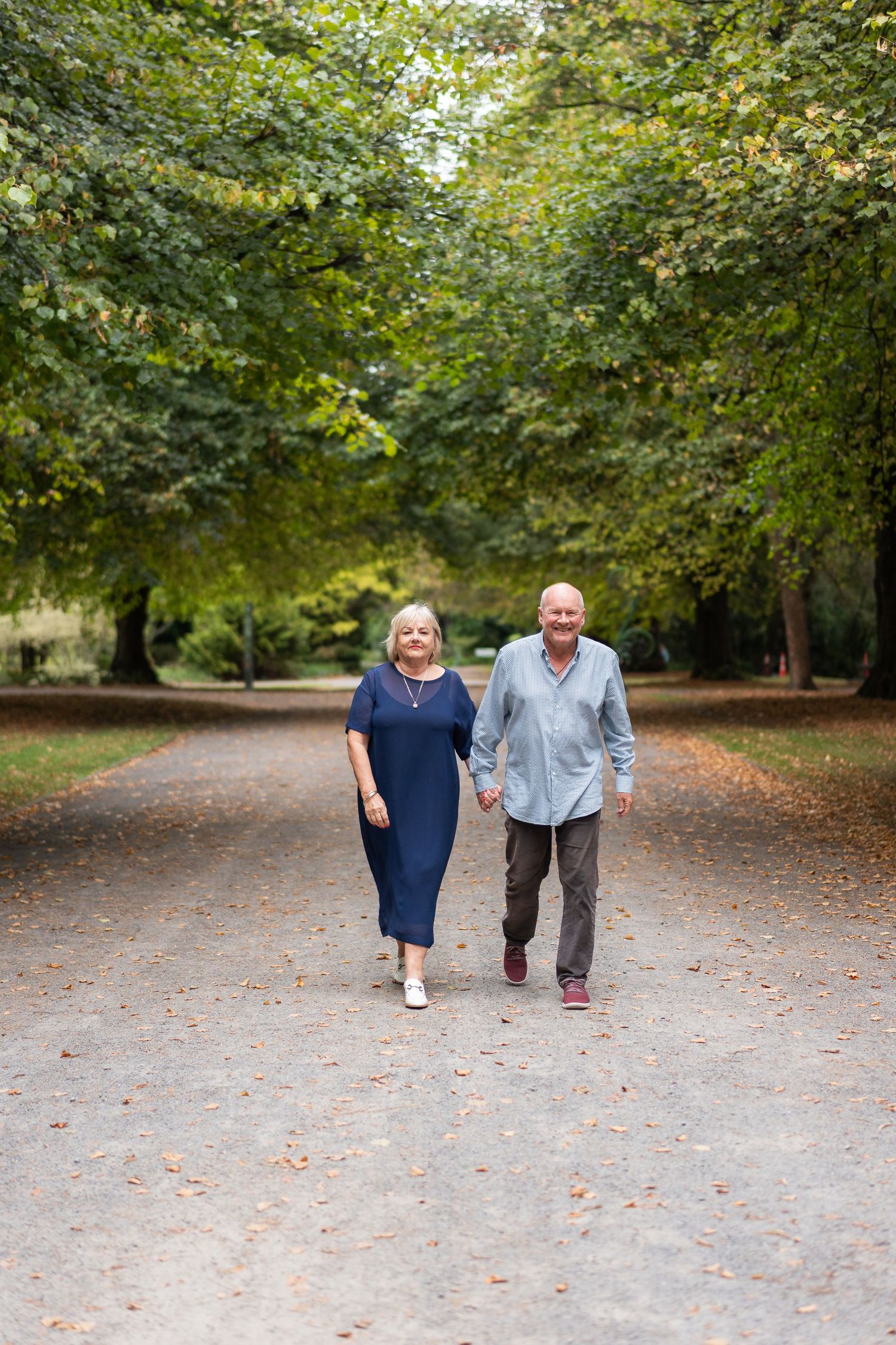 Elderly couple holding hands and walking together on a tree-lined park path, smiling warmly on a sunny day.