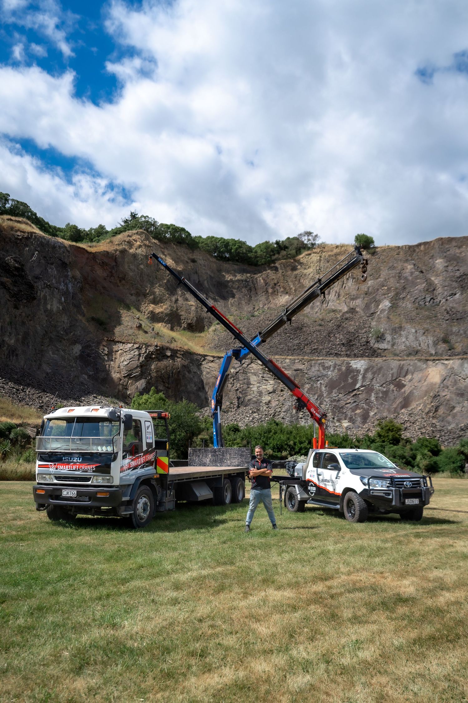 A crane truck and white ute parked on grass near a quarry face, with a worker standing between the vehicles.