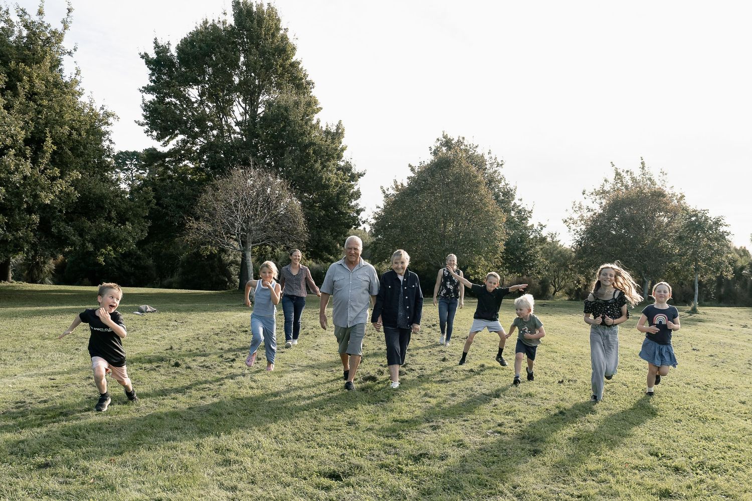 Multi-generational family running and playing together on a sunny green park lawn surrounded by tall trees.