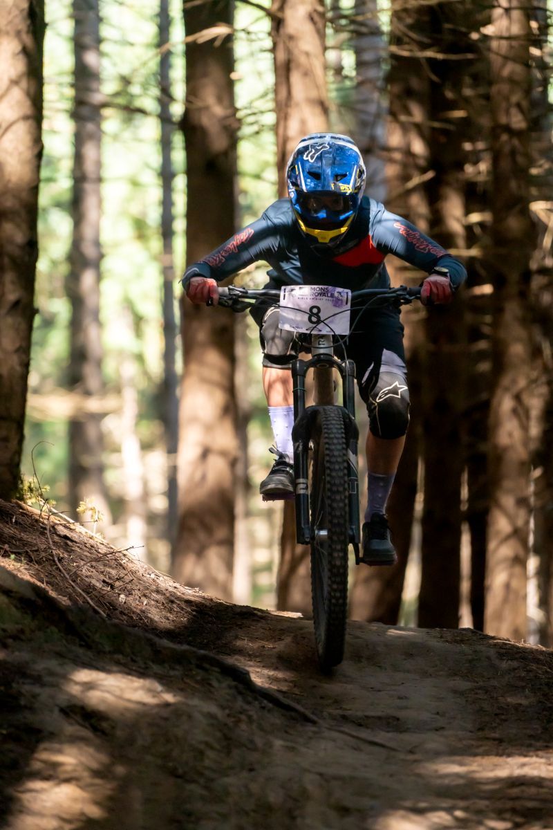 Mountain biker descending steep forest trail wearing protective gear and helmet during downhill ride through pine trees.