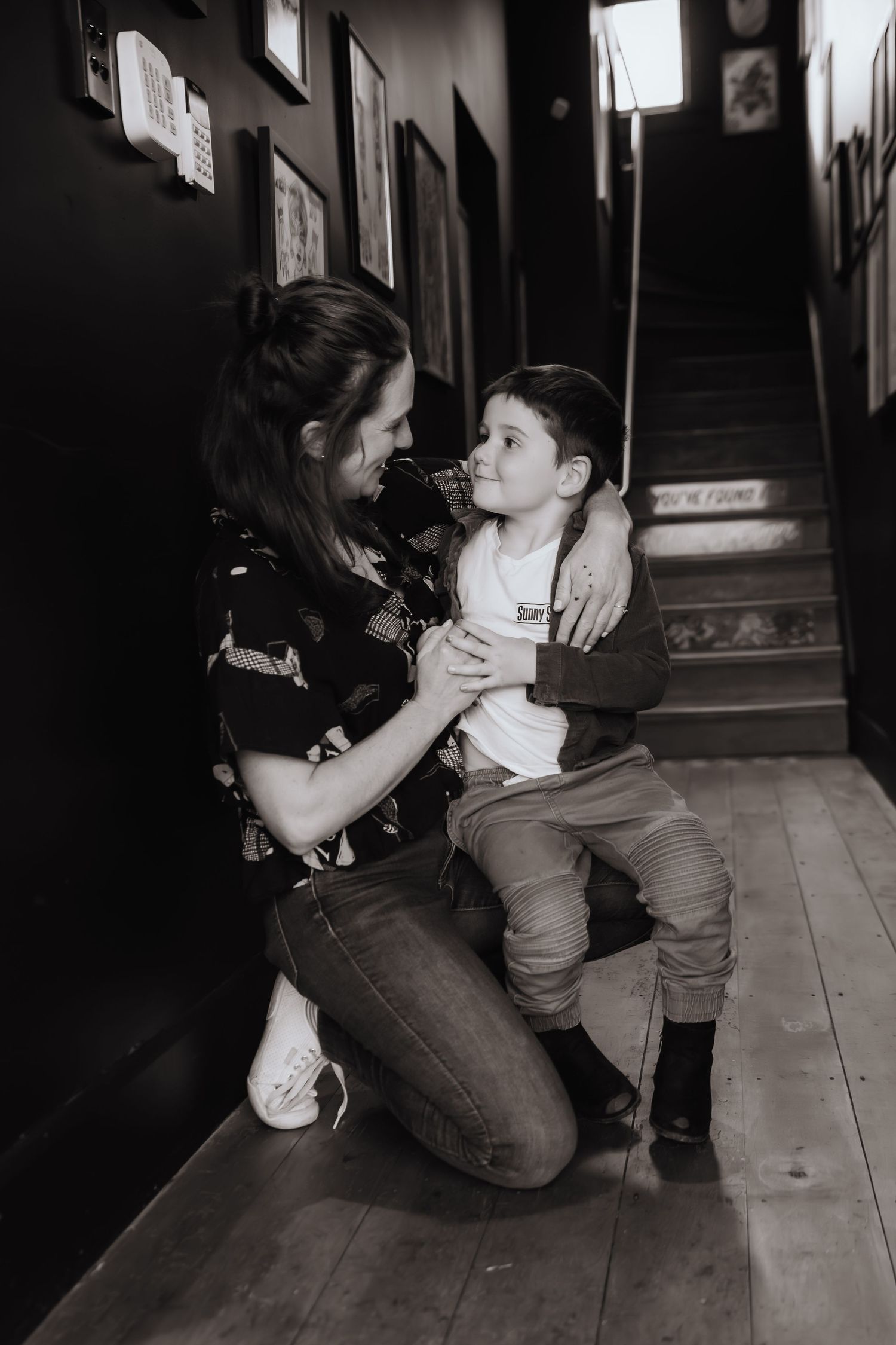 Black and white photo of a mother kneeling to embrace her young son in a warmly decorated hallway.