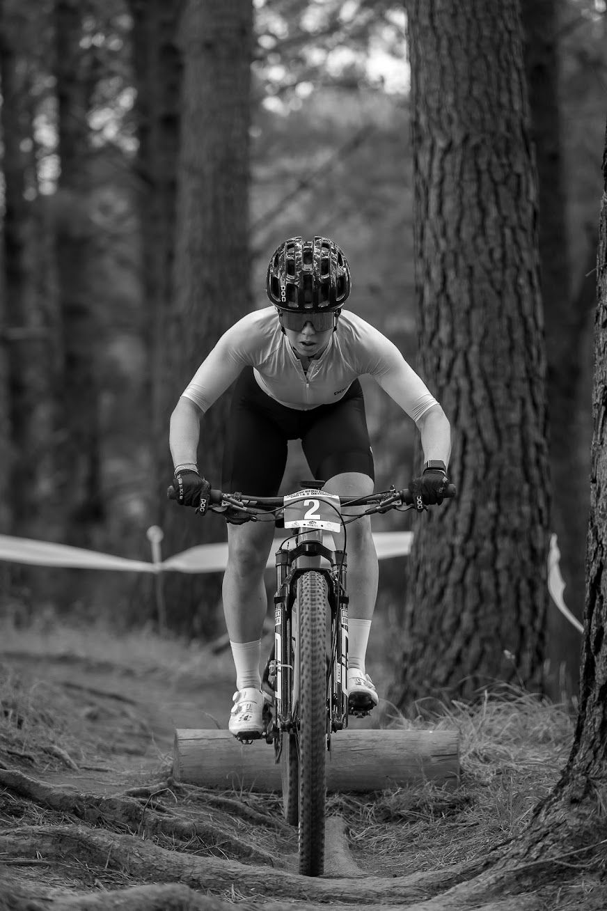 Black and white photo of a mountain biker racing through a forest trail wearing a helmet during a competition.