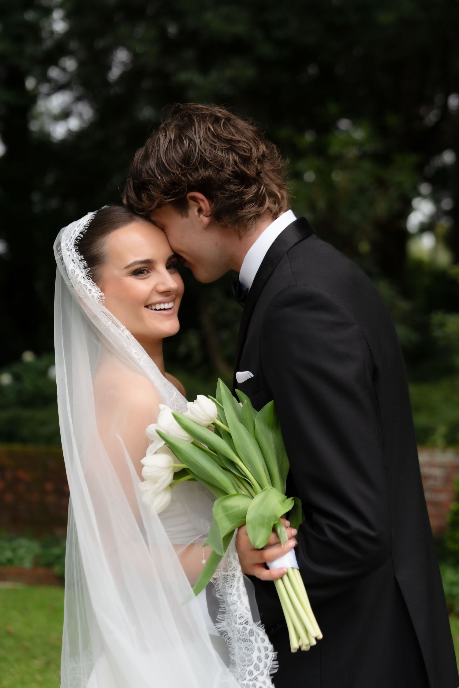 Bride in white veil holds tulip bouquet as groom in black tuxedo leans in for a kiss outdoors.
