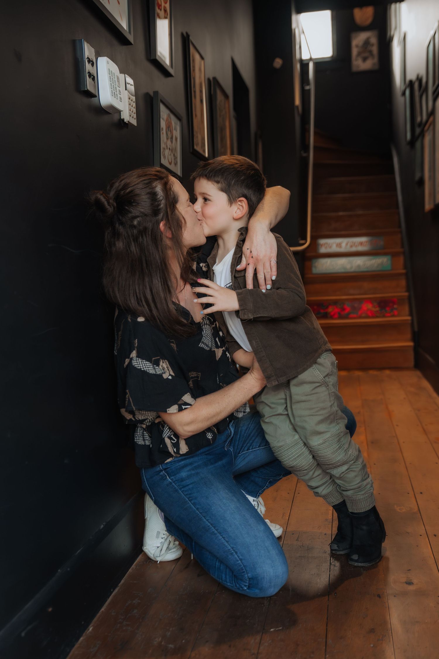 A mother kneeling on hardwood floor hugging and kissing her young son in a dark hallway with stairs.