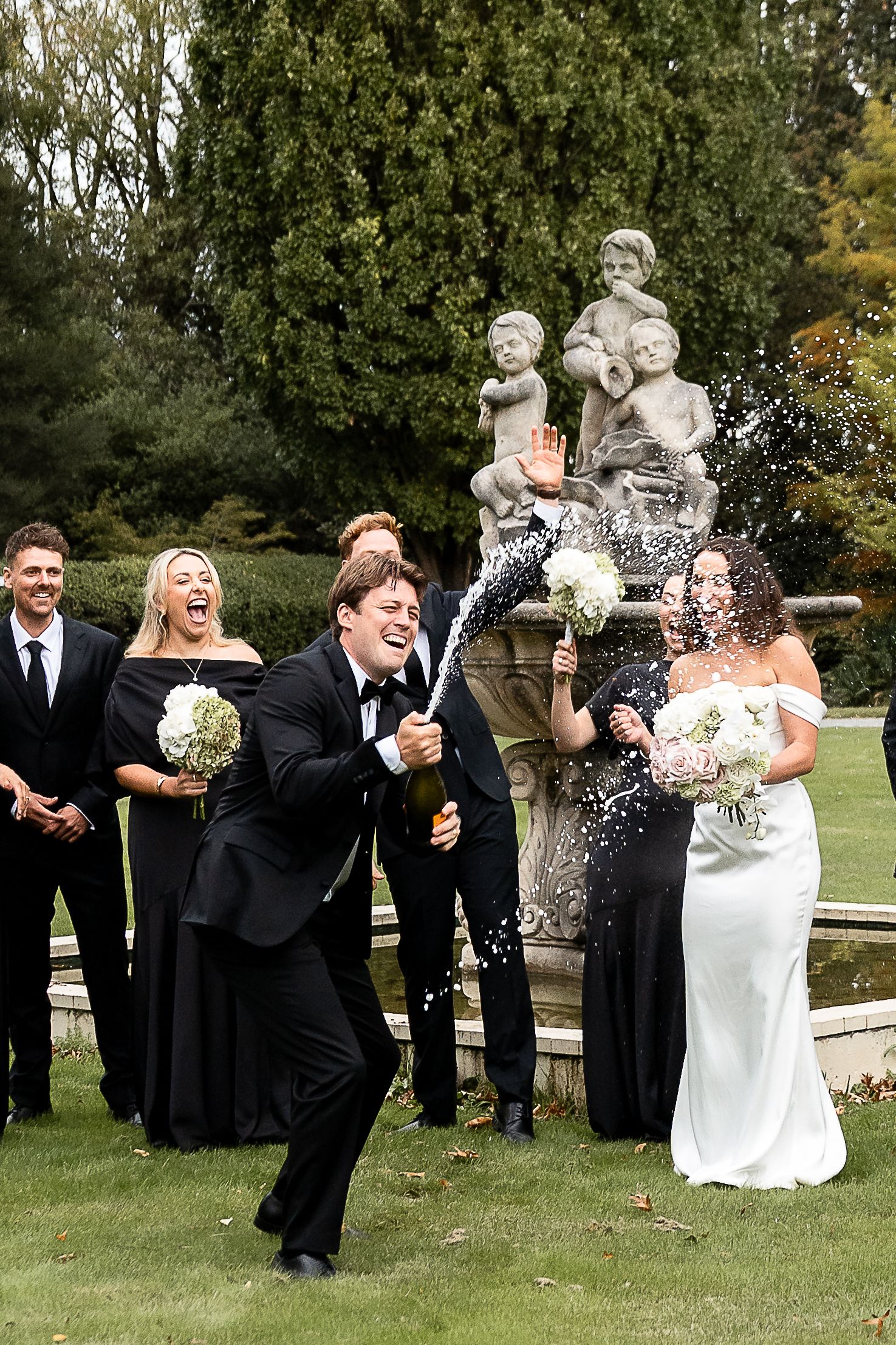 Bride and groom celebrate with champagne spray at outdoor wedding, bridal party in black laughing behind stone fountain.