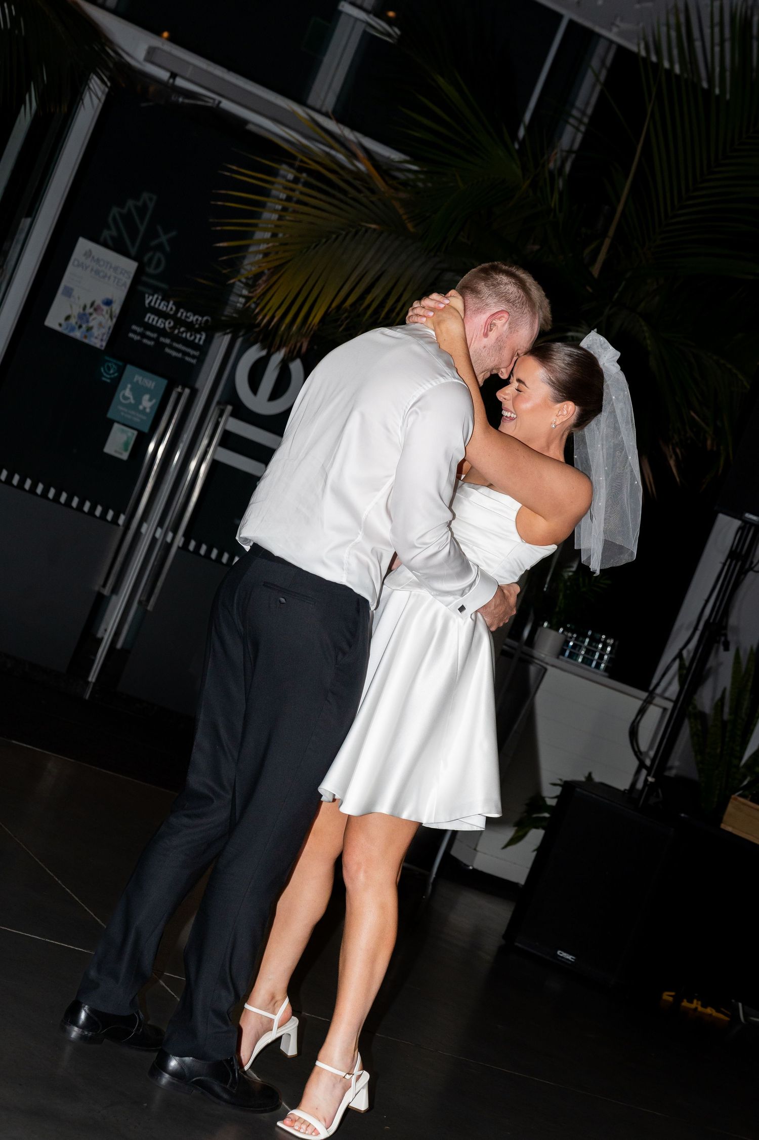 Bride and groom sharing their first dance, bride in short white dress and veil, groom in white shirt and black pants.