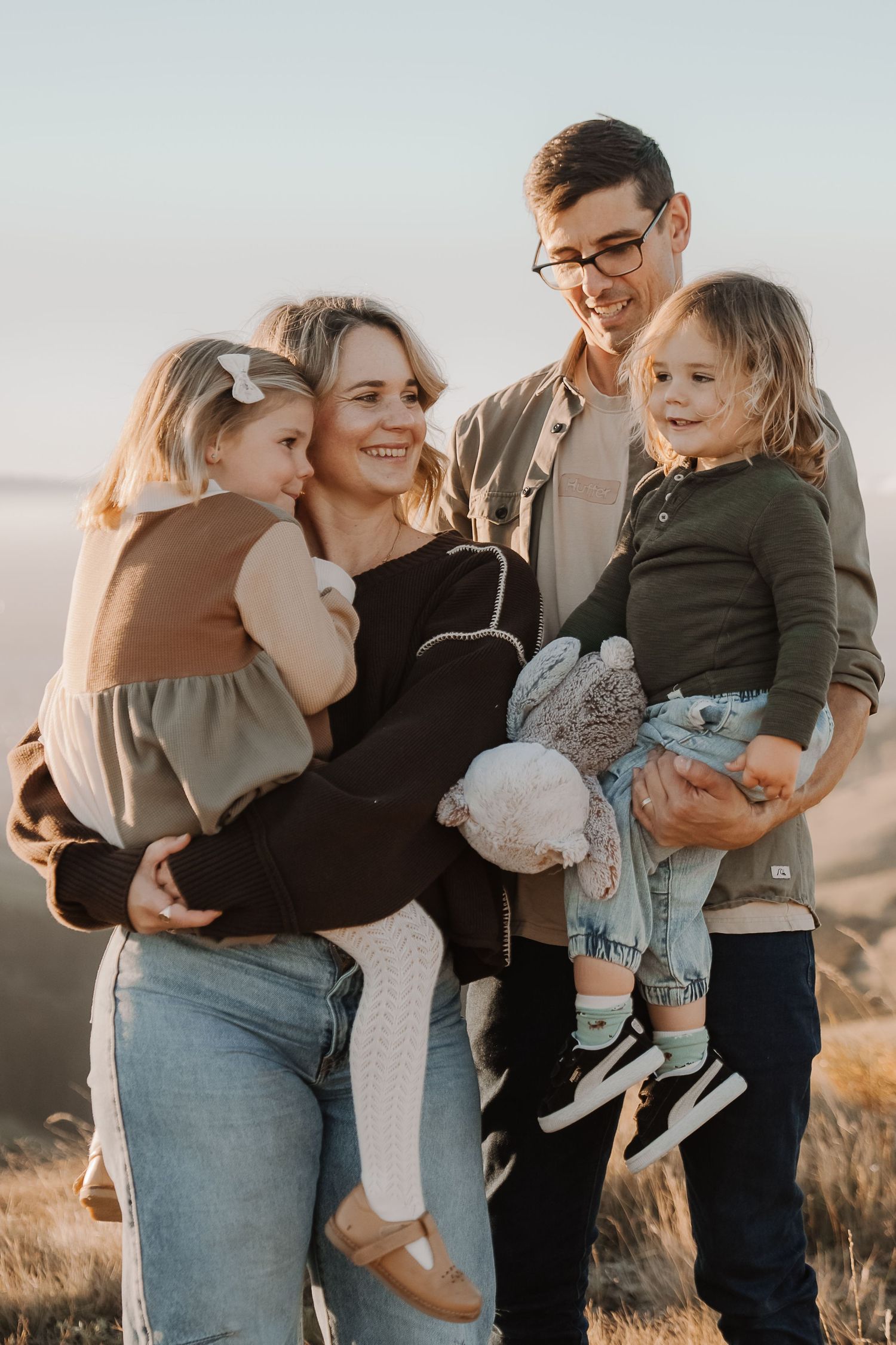 A smiling family of four poses outdoors at sunset, parents holding two young children in a golden grassy field.