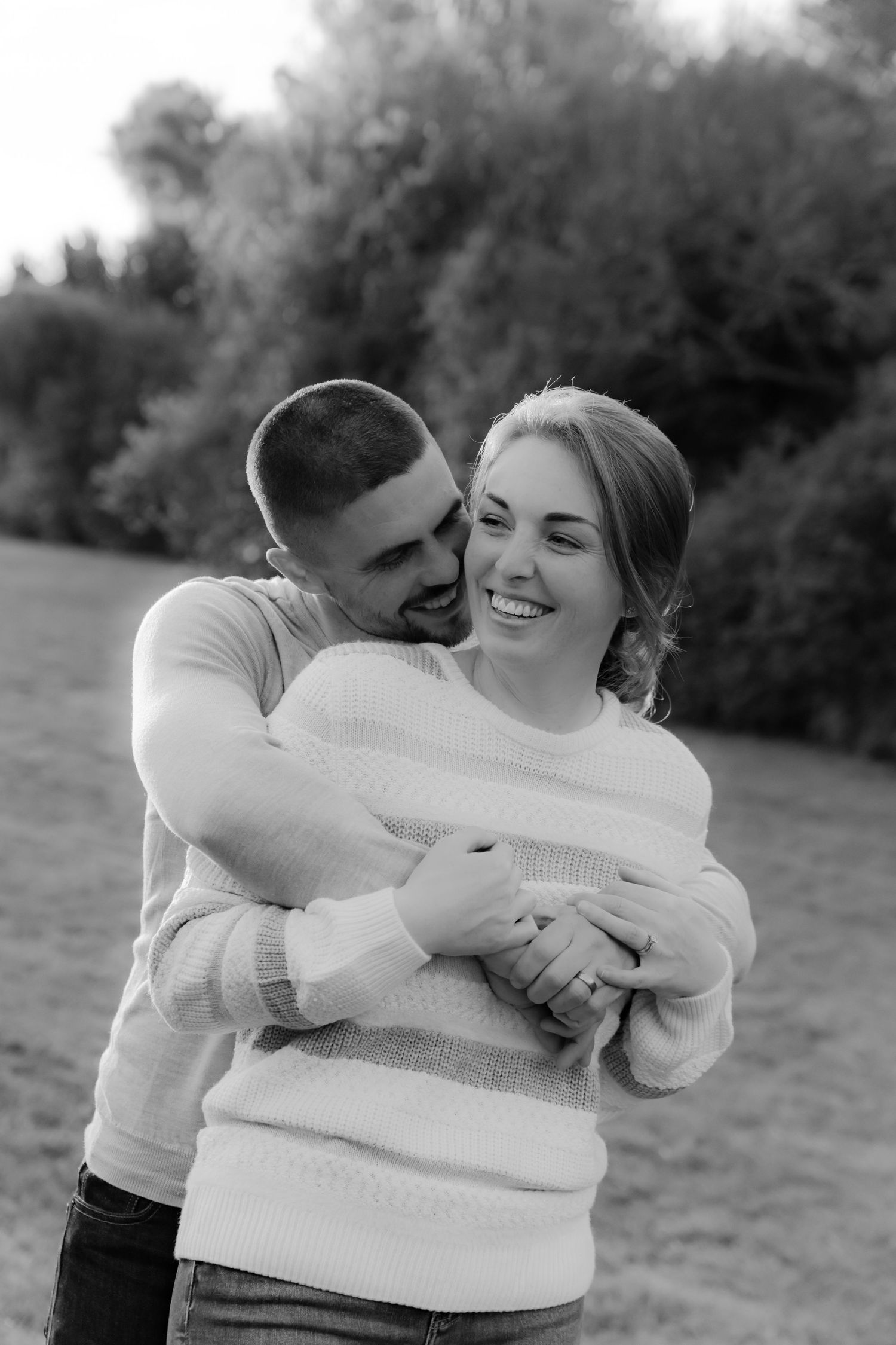 Black and white photo of a couple embracing outdoors, man hugging woman from behind as she smiles joyfully.