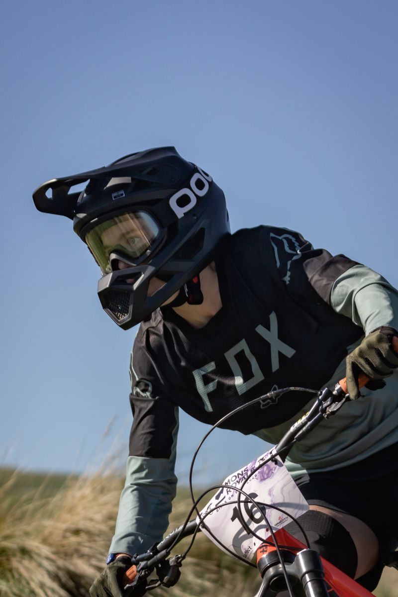 Mountain biker wearing protective gear and Fox apparel rides downhill against blue sky background.