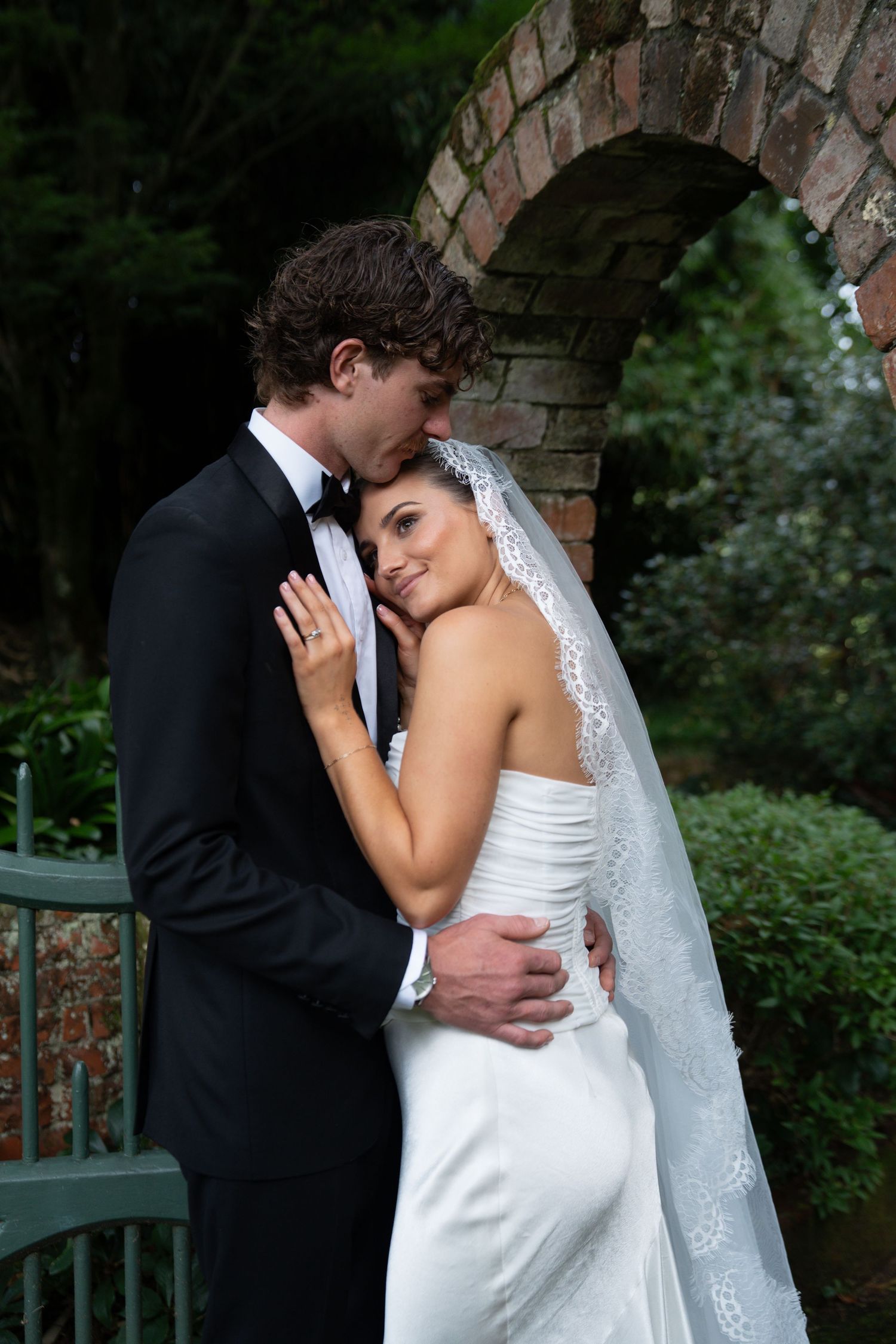 A bride and groom embrace tenderly in a garden setting, she in a white gown and veil, he in a black tuxedo.