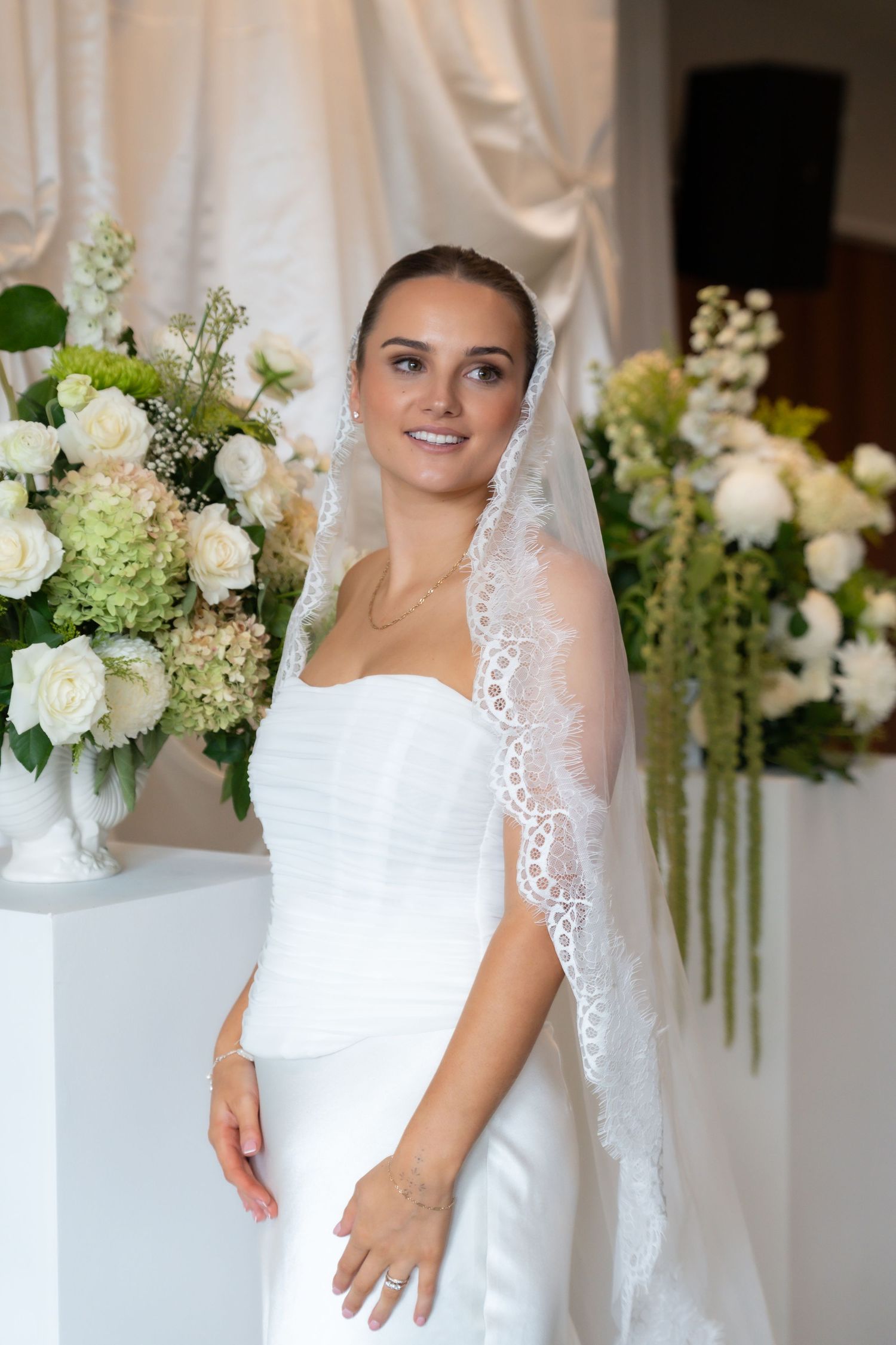 New Zealand bride in white strapless gown and lace veil smiling, surrounded by elegant white floral arrangements.