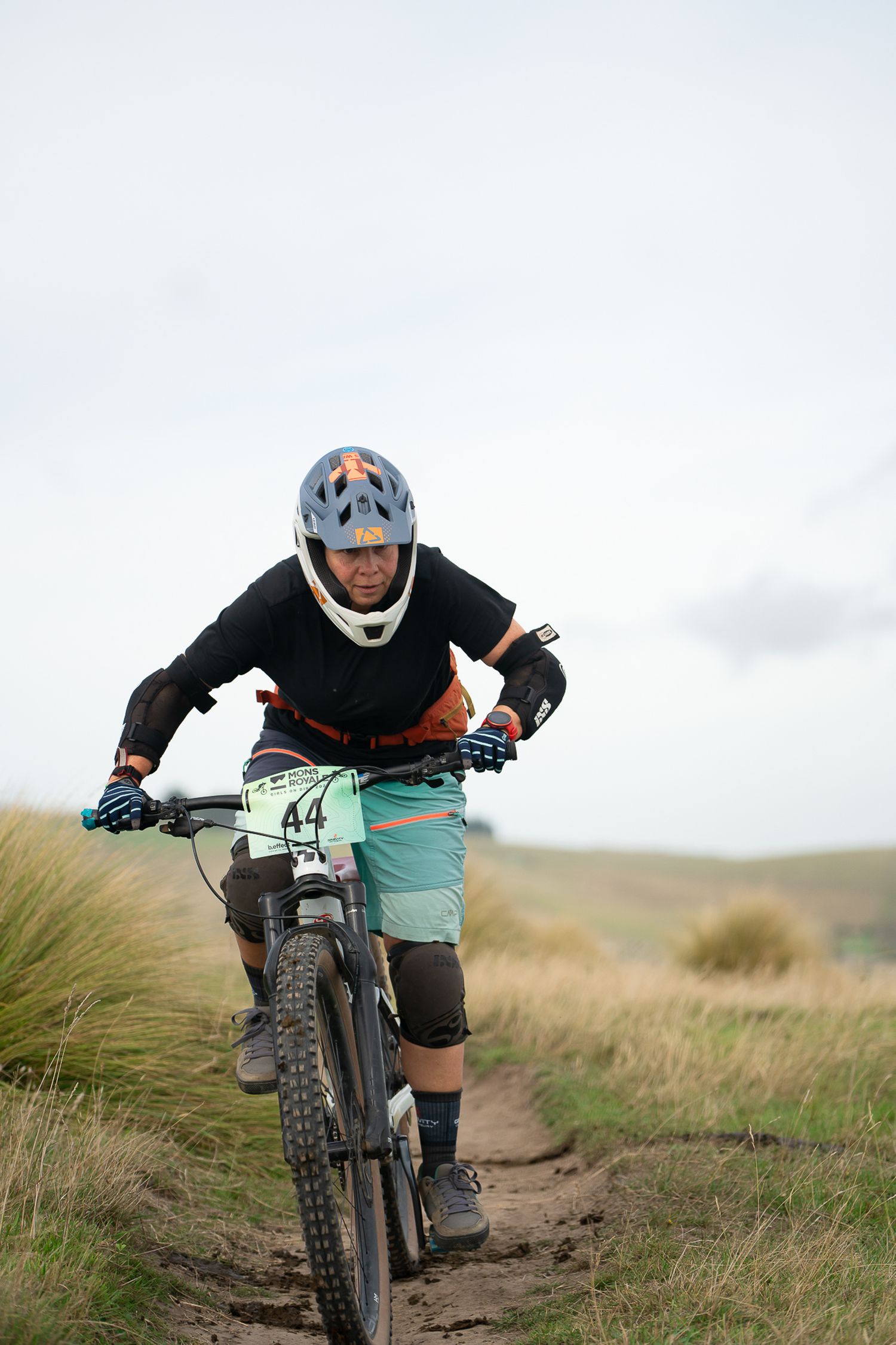 Female mountain biker in protective gear navigates a dirt trail through grassy terrain on a cloudy day at the Crocodile Mountain Bike Park.