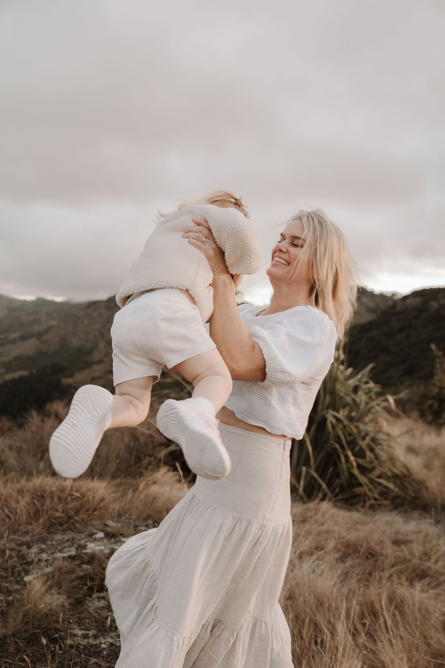 Smiling blonde woman lifting a happy toddler in white outfit against scenic mountain backdrop outdoors.