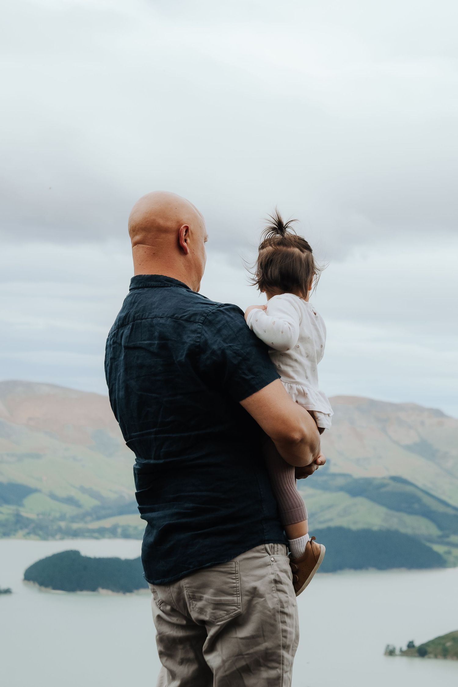 Bald man holding a young child overlooking a scenic harbor with green hills under a cloudy sky.