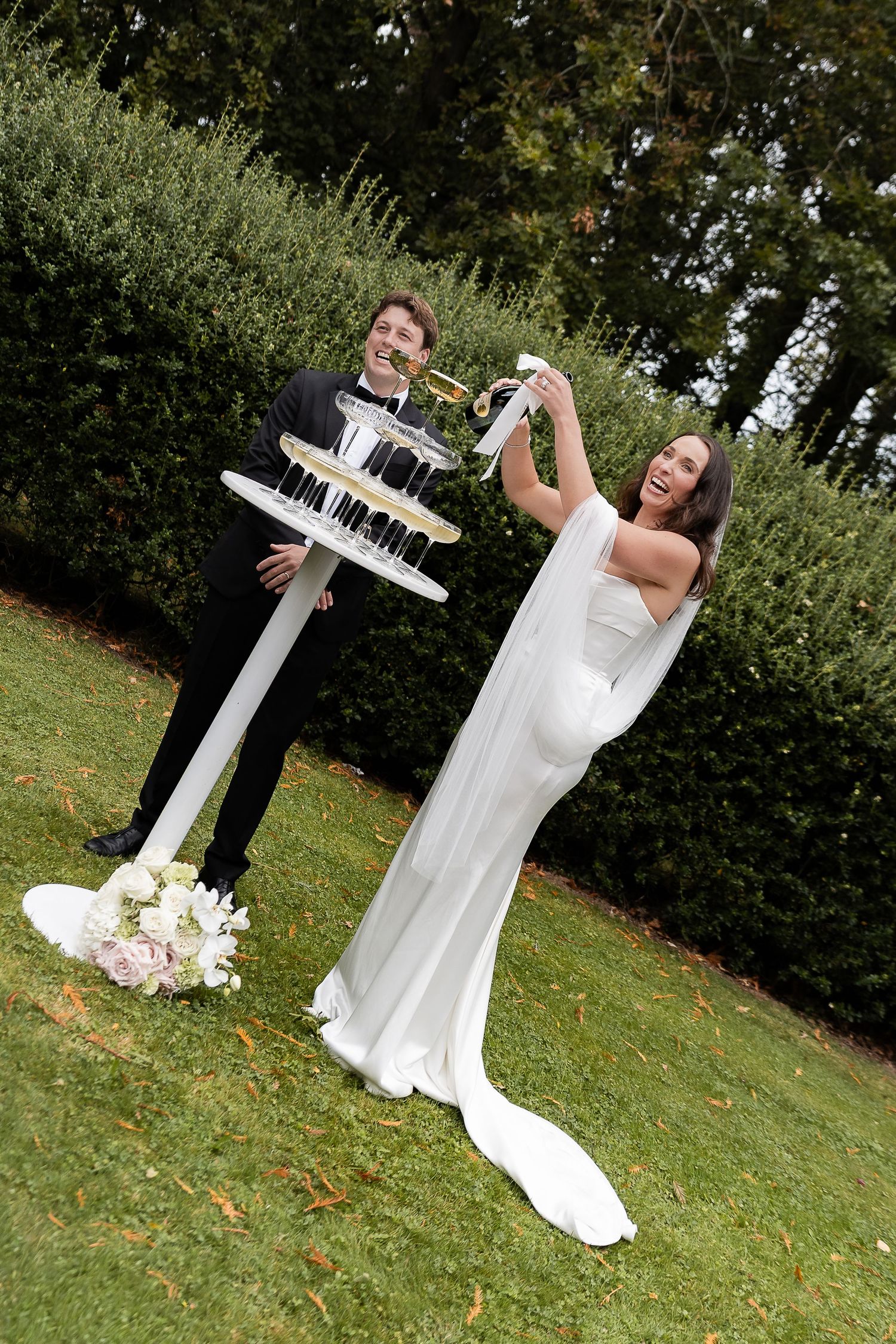 Bride and groom celebrating outdoors with champagne tower on green lawn, bride in elegant white gown laughing joyfully.