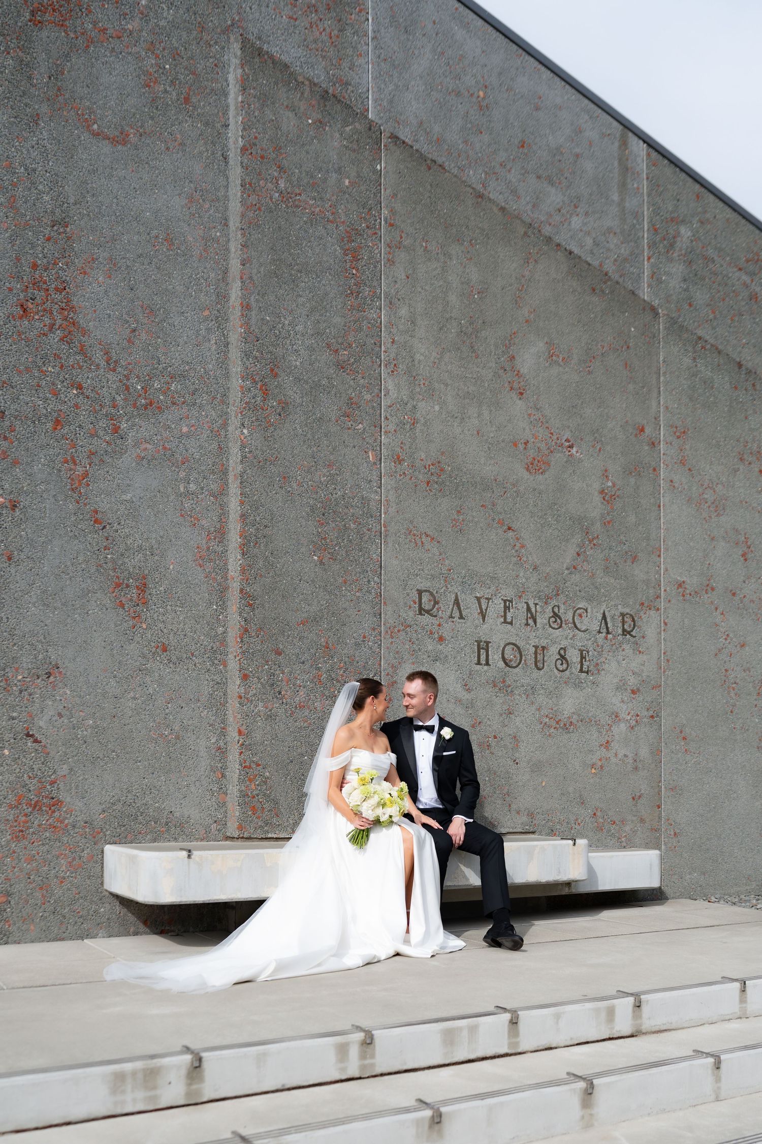 Christchurch bride and groom sit together on stone steps outside Ravenscar House, bride holding white bouquet in wedding dress and veil.