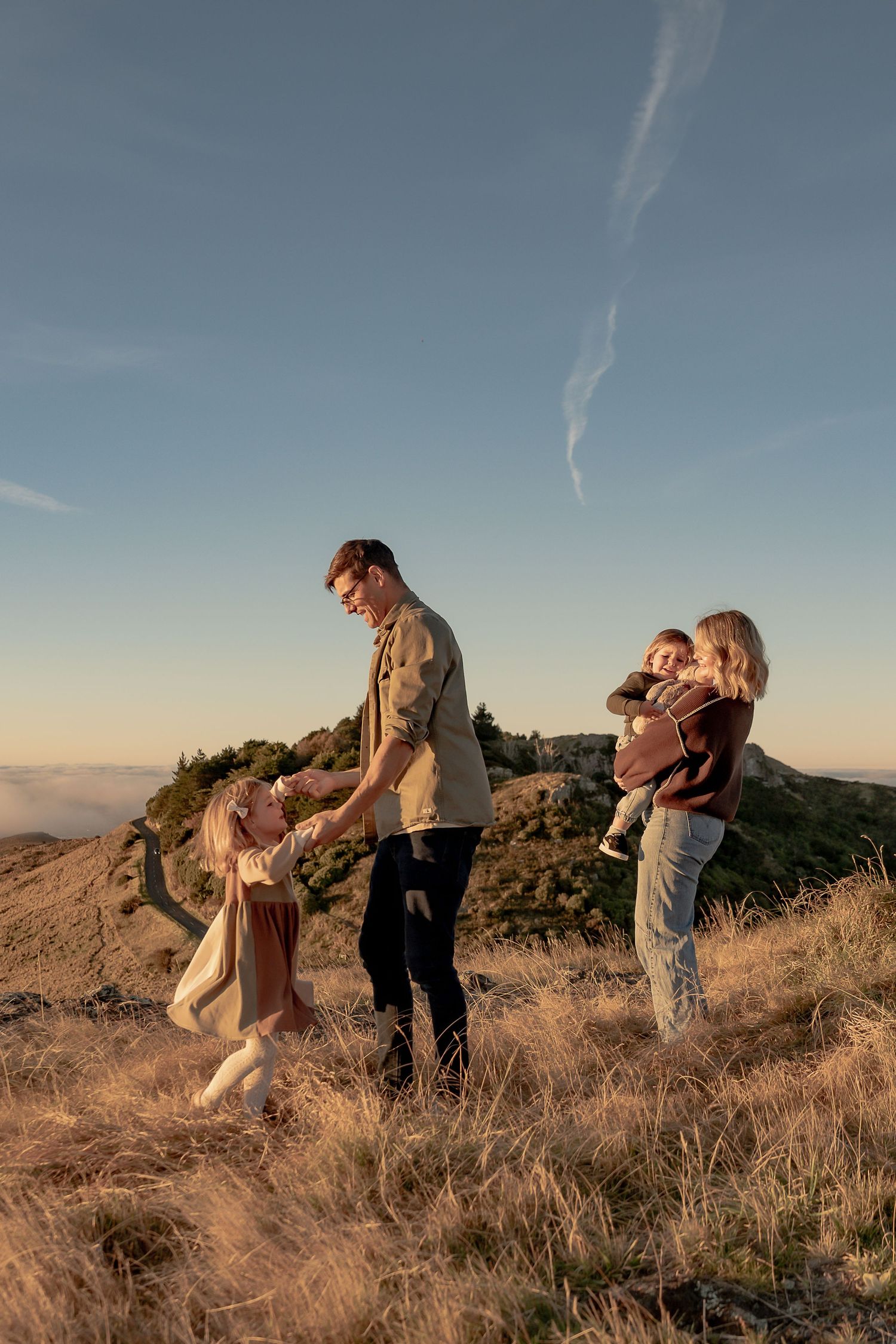 A family of four enjoys a golden hour sunset on a grassy hilltop, with parents playing with two young children.