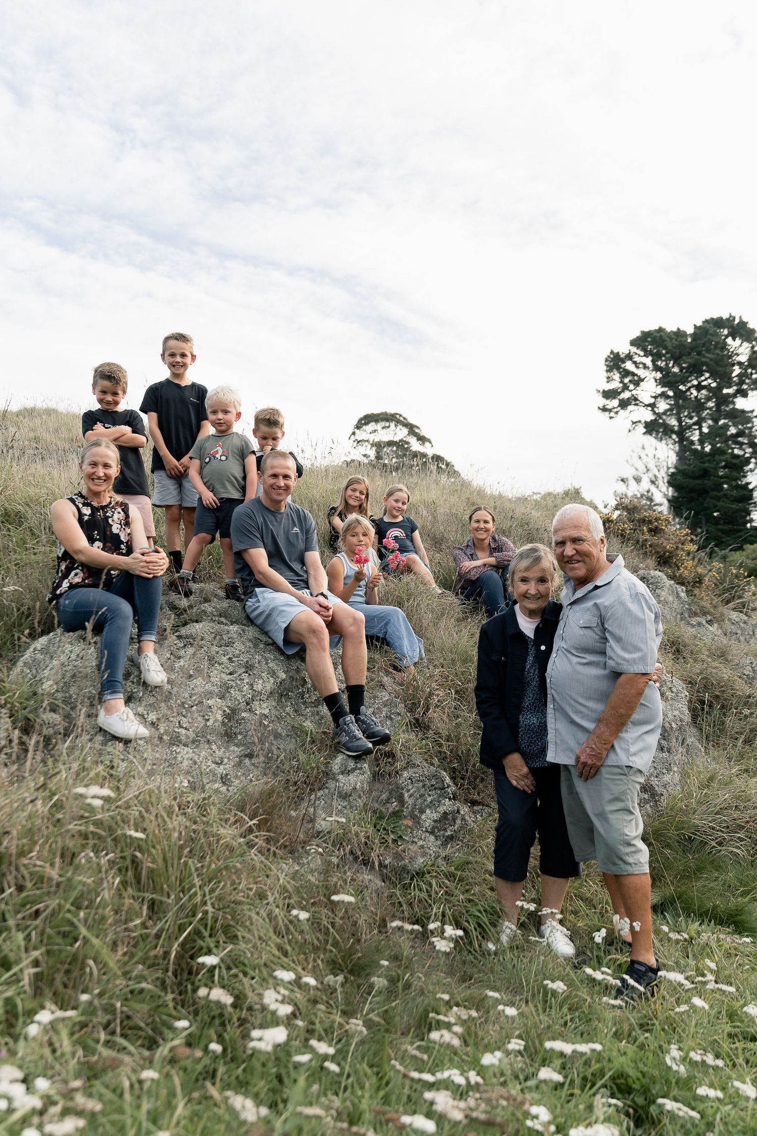 Large family group posing on rocky outcrop in grassy field, smiling for outdoor portrait on cloudy day.