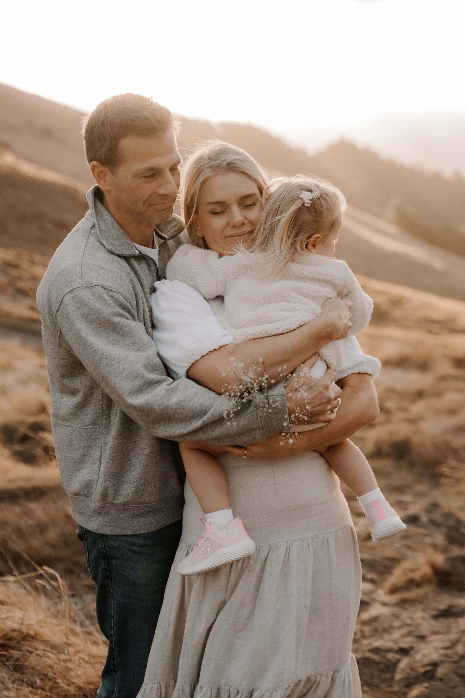 A loving family of three shares a warm embrace outdoors at golden hour, with parents holding their toddler.