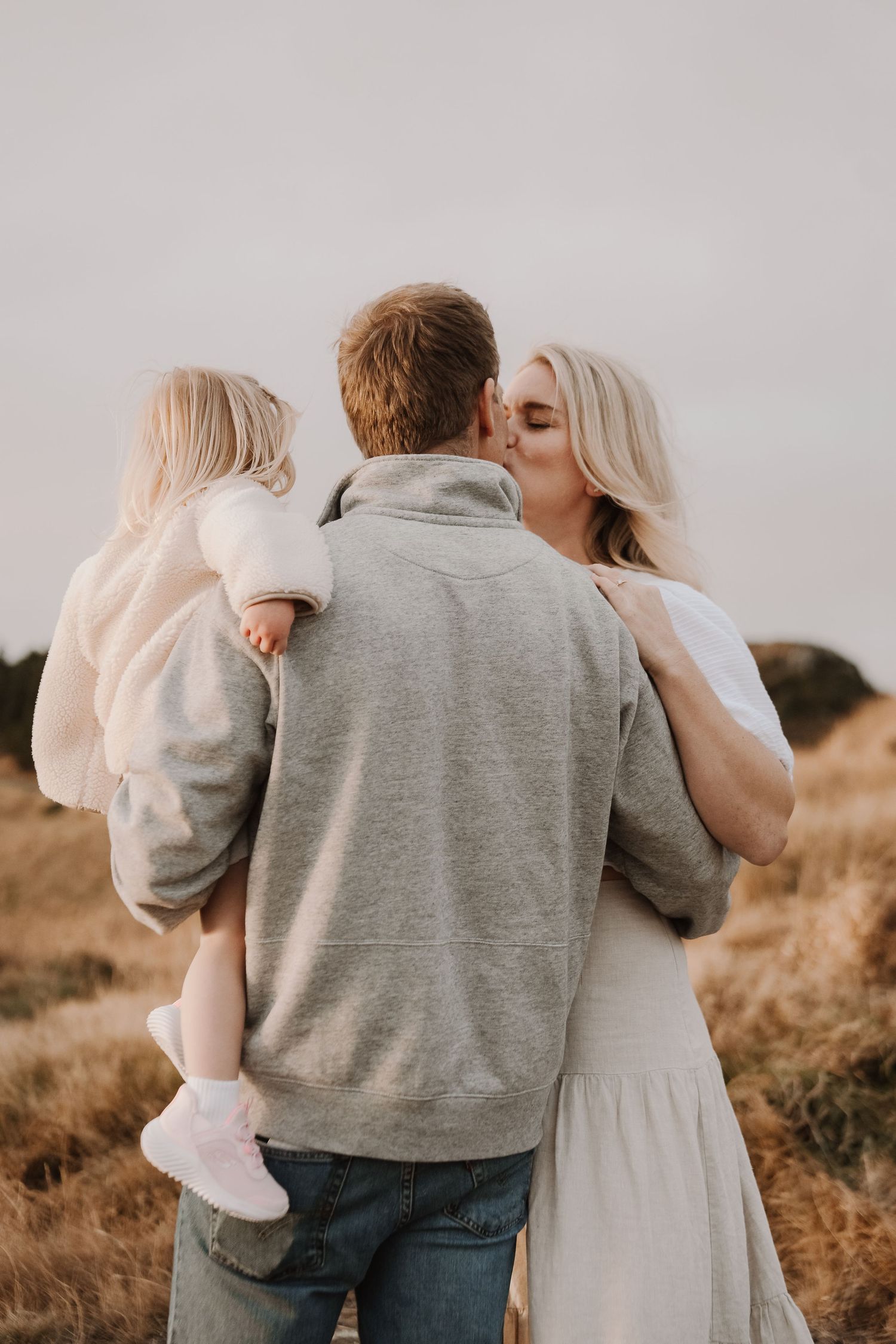 A couple shares a kiss outdoors while holding their young blonde toddler in a golden field setting.