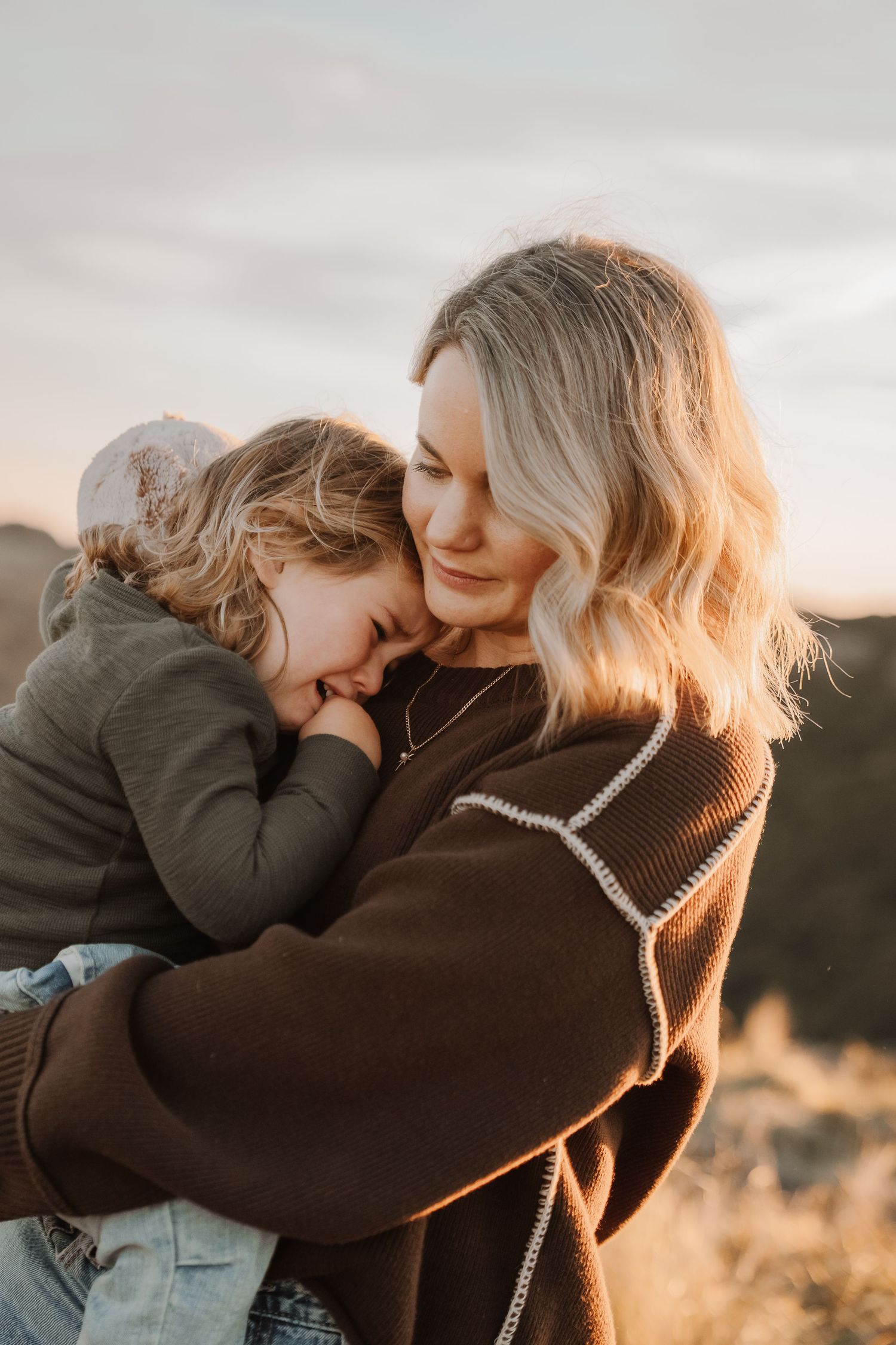 Mother tenderly holds young child in warm embrace during golden hour outdoor photography session.