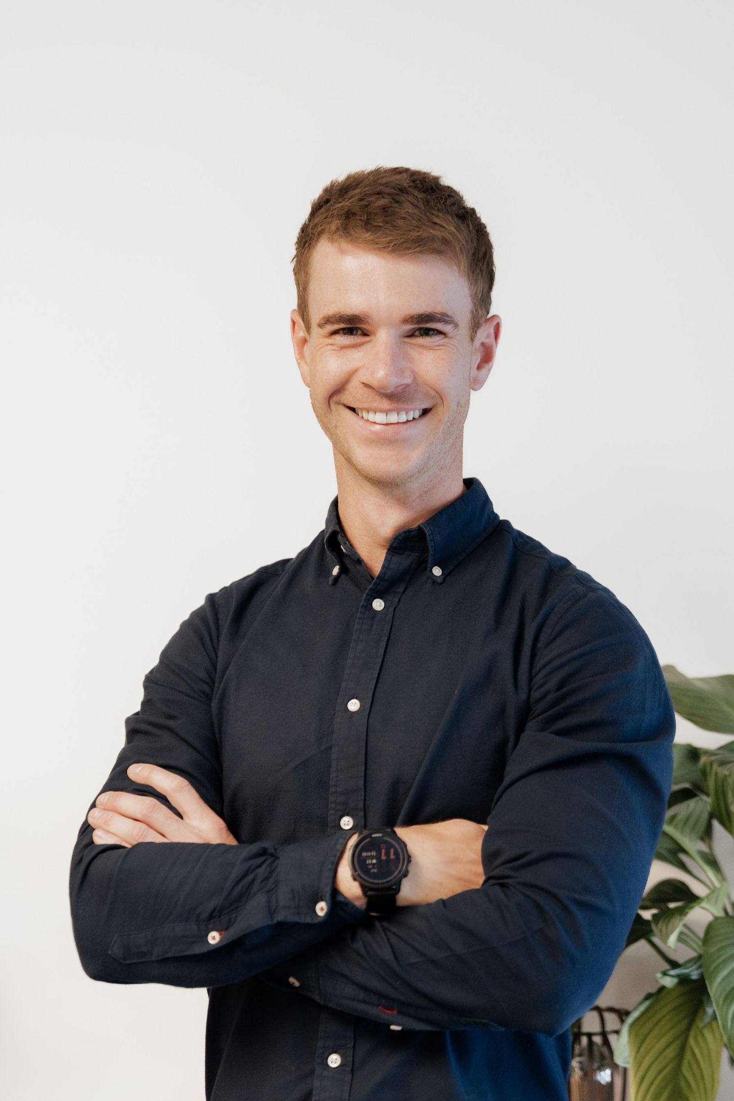 Smiling young man in dark navy shirt with arms crossed, wearing a black watch, posing against a white background.