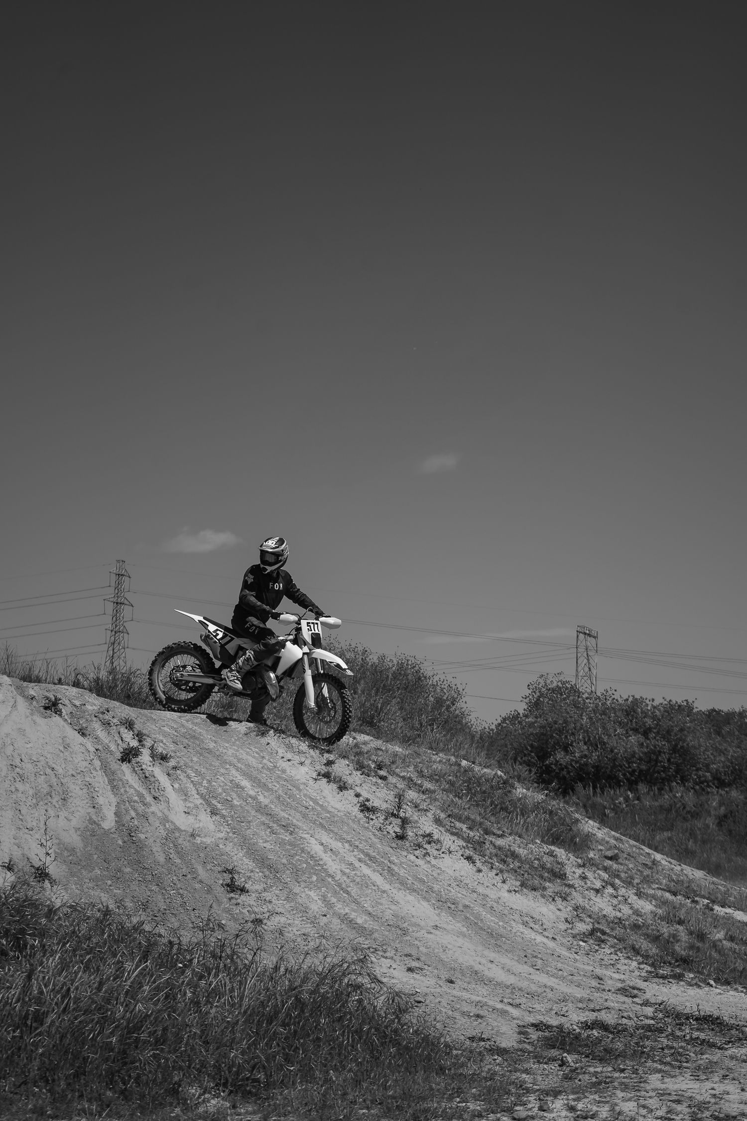 A motocross rider about to ride down a steep dirt hill against a cloudy sky with power lines visible in the background in Christchurch.