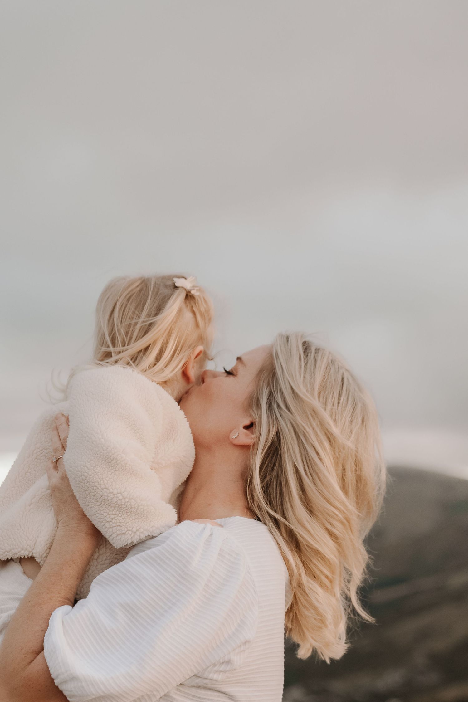 A blonde mother lovingly kisses her young toddler daughter outdoors against a soft gray sky.
