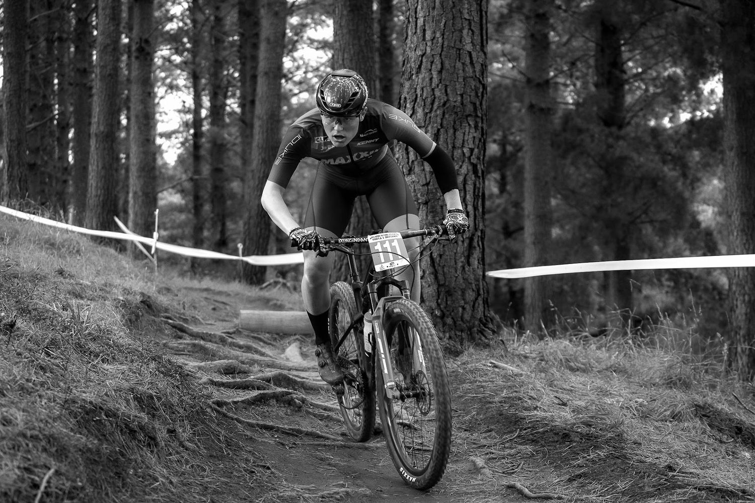 Black and white photo of a mountain biker racing downhill through a forest trail during a competitive cycling event.