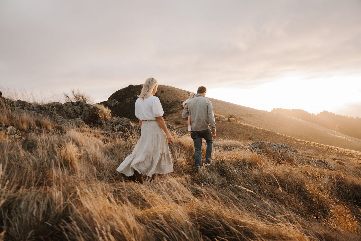 Couple walking through golden grass fields at sunset on a scenic hillside, woman in white dress.