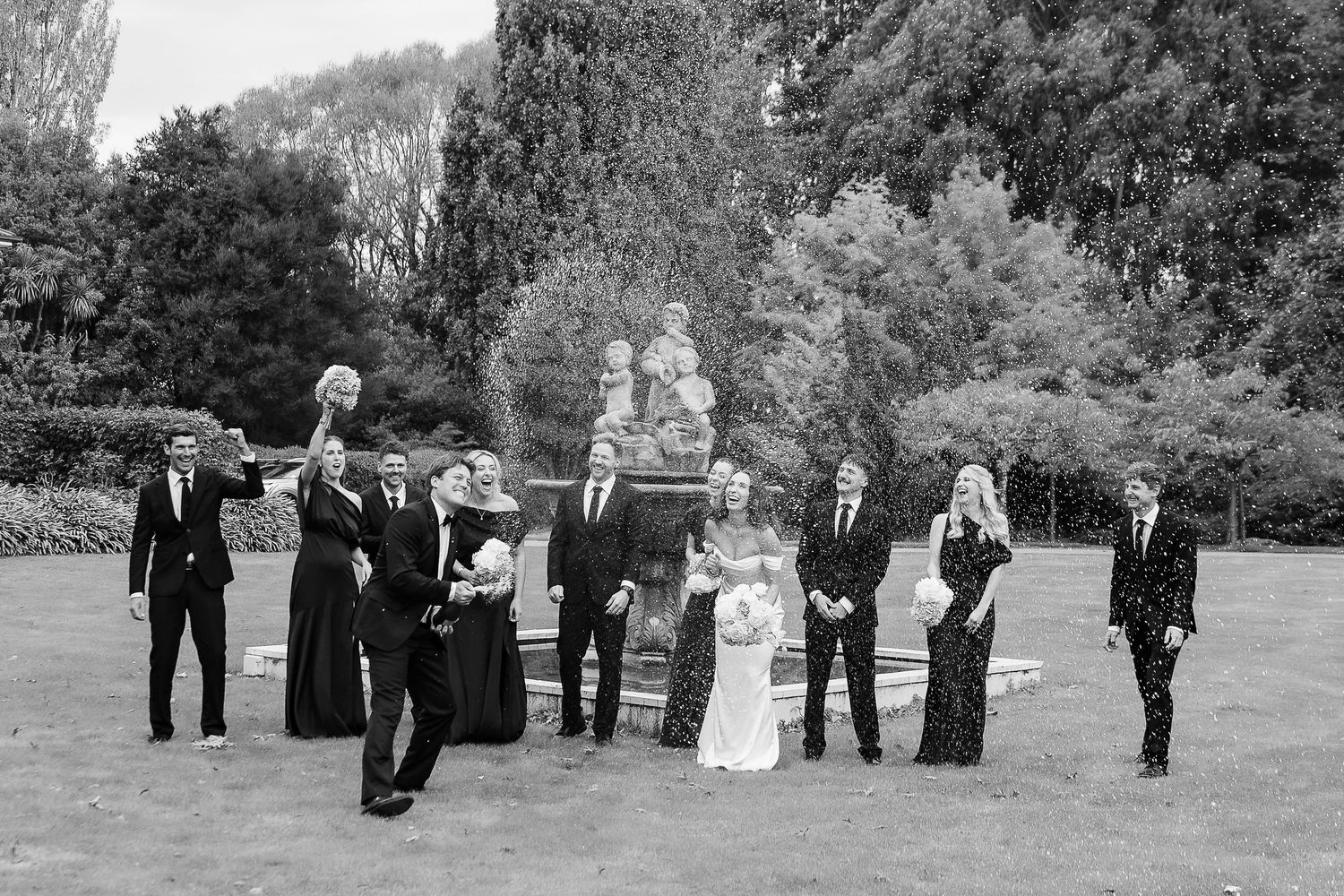 Black and white wedding party photo outdoors near stone fountain, bridal party celebrating in formal attire.